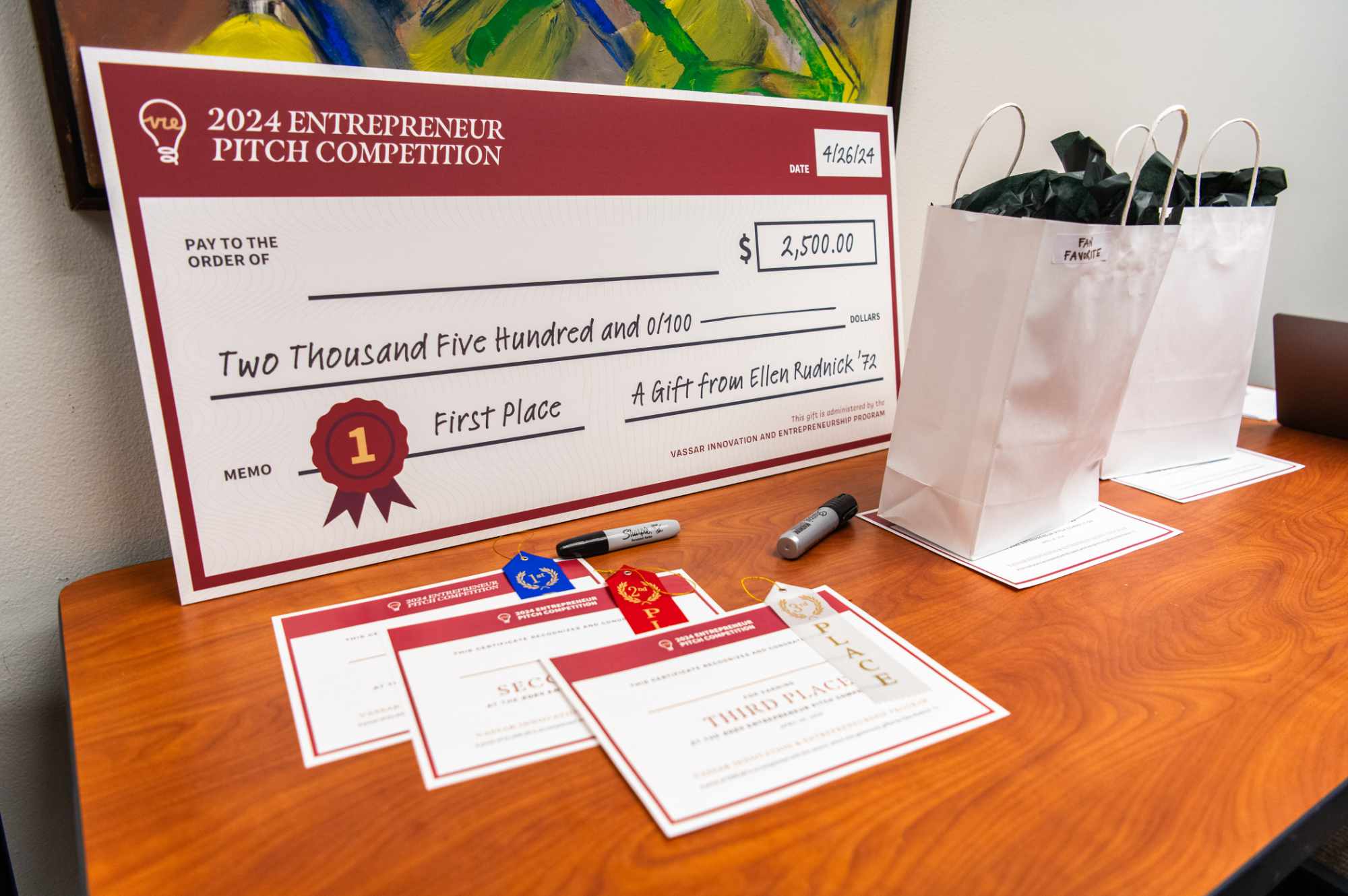 A large poster-sized check on a wooden table, with signup forms near it. Printed on the check is the text “2024 Entrepreneur Pitch Competition. First Place, Two Thousand Five Hundred Dollars. A Gift From Ellen Rudnick ’72”.