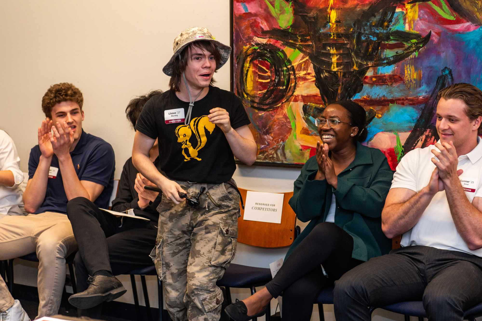 Three people sitting in chairs, clapping, looking at a fourth standing person in front of them who appears to be speaking or performing.