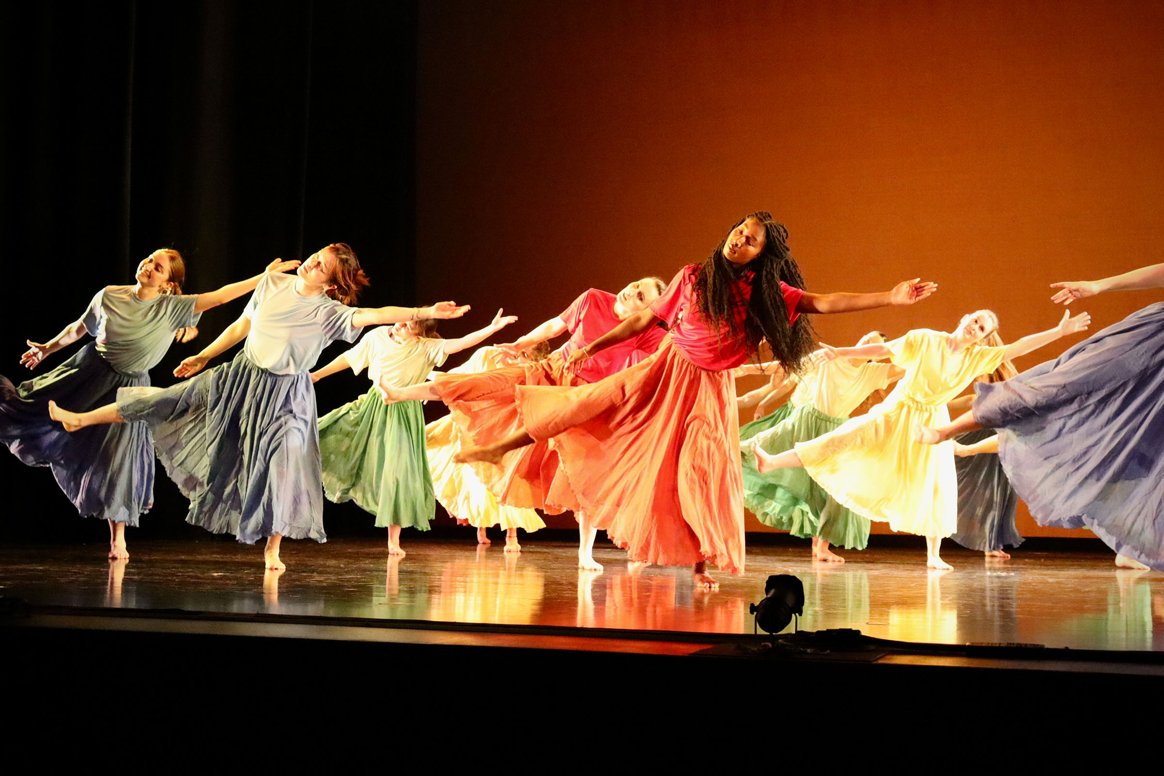 A large group of people dancing on a stage. They are all wearing different color dresses while leaning over on one leg with their arms out straight in unison.