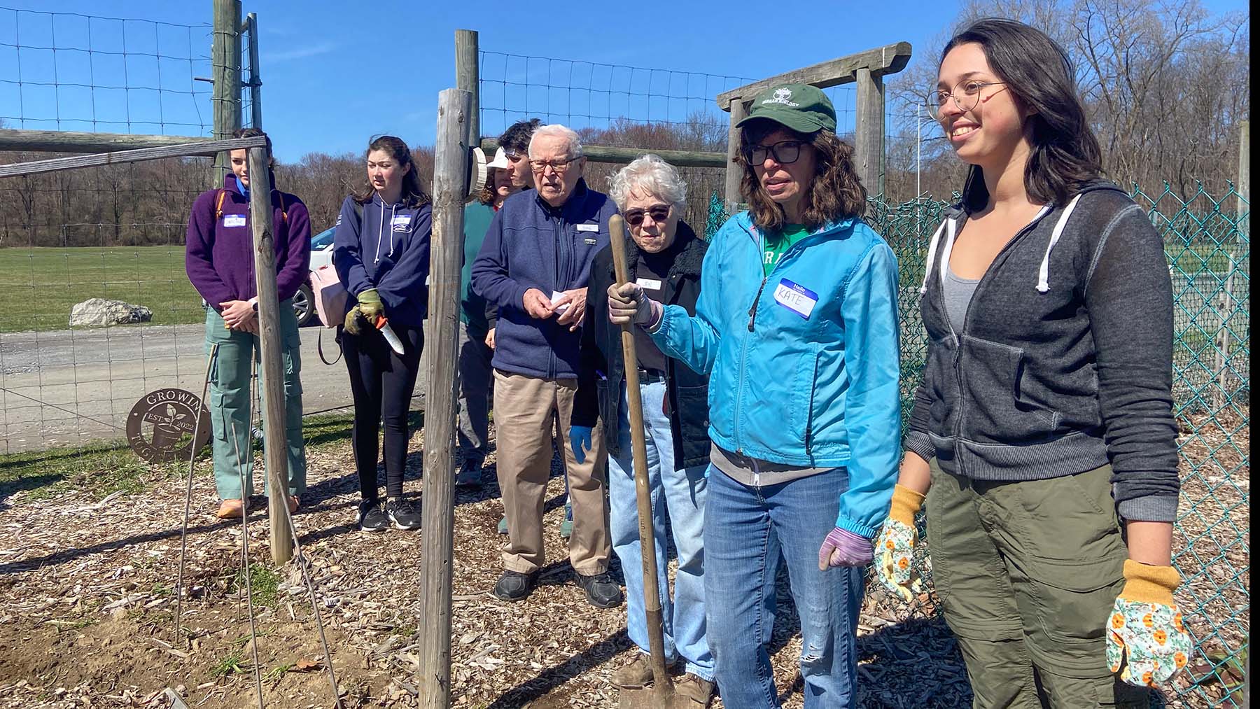 A group of adults of various ages stand by a fence in a sunny field.