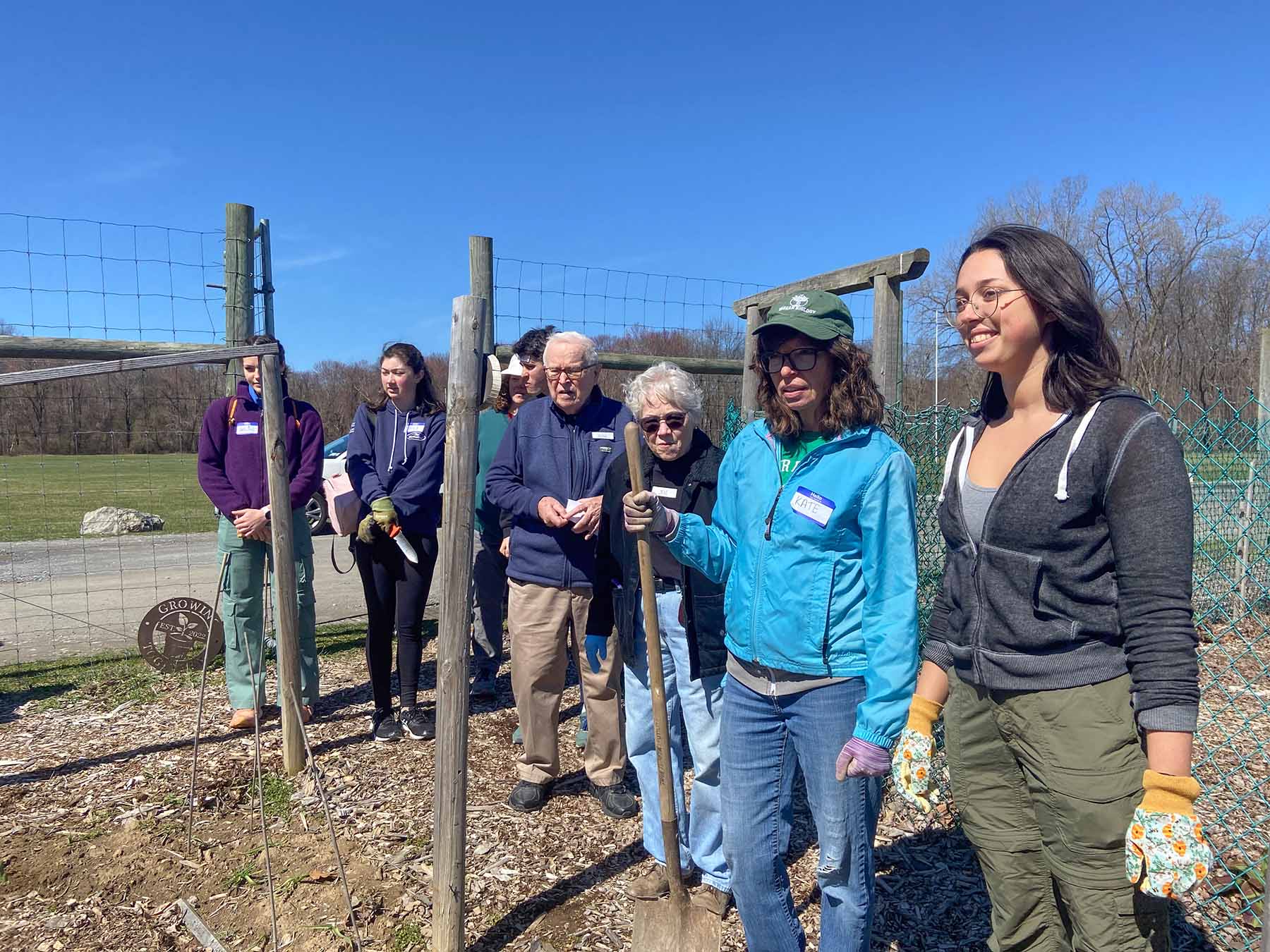 A group of adults of various ages stand by a fence in a sunny field.