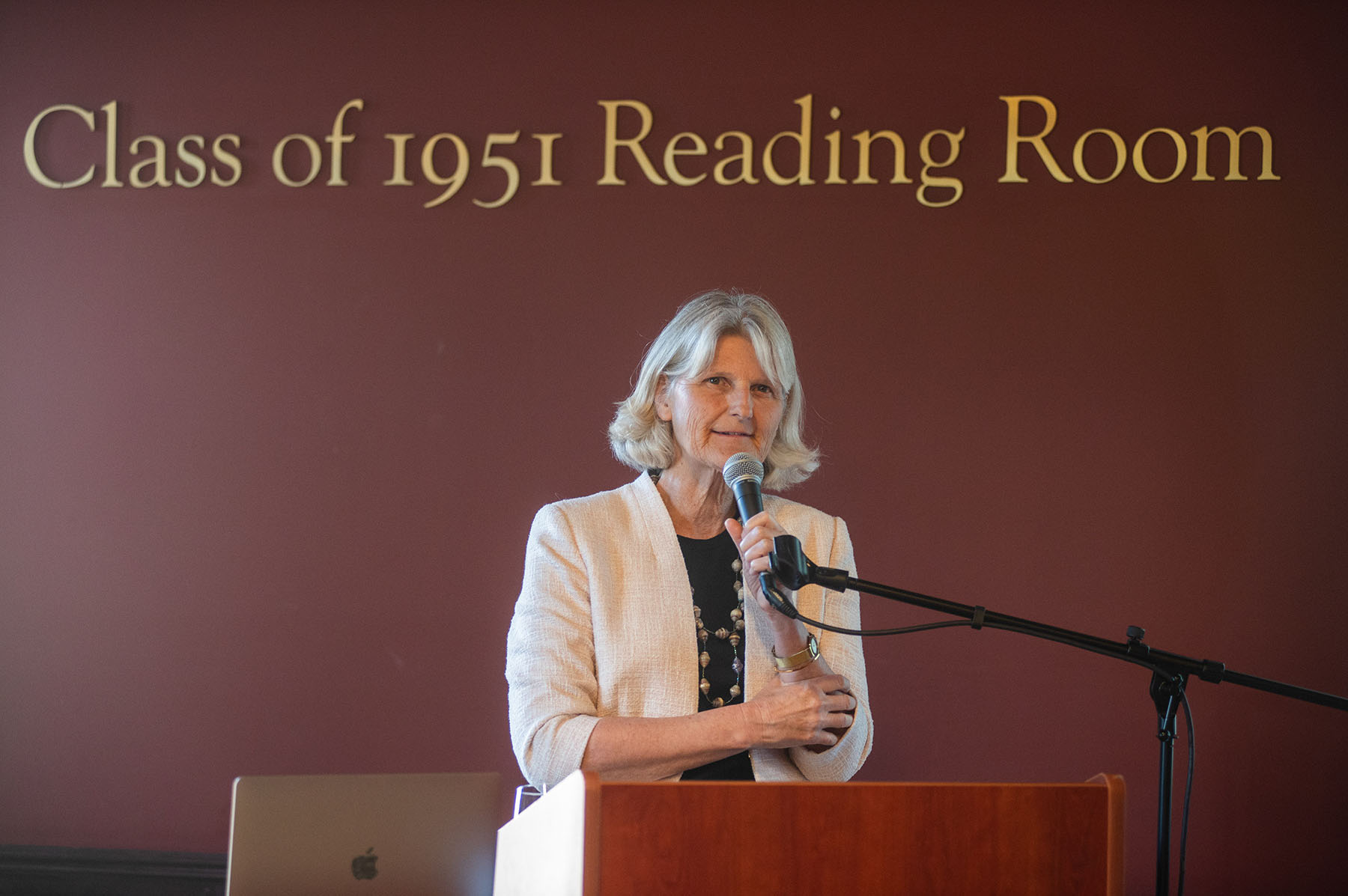 President Elizabeth H. Bradley, wearing a black shirt and white jacket, speaking at podium in front of 1941 reading room sign.