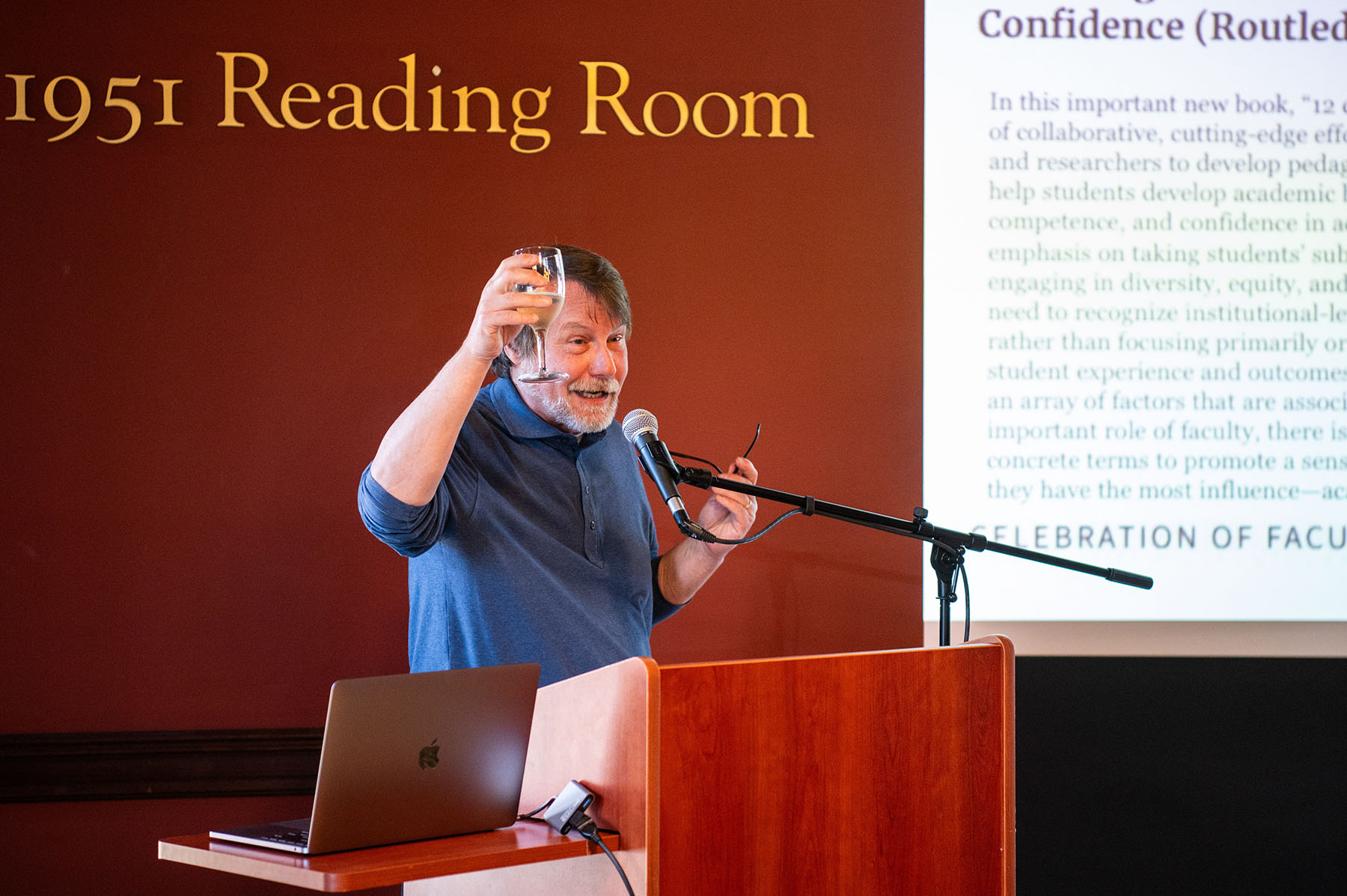 Dean of Faculty William Hoynes standing at a podium using a projector screen for his presentation, his hand in the air holding a glass.