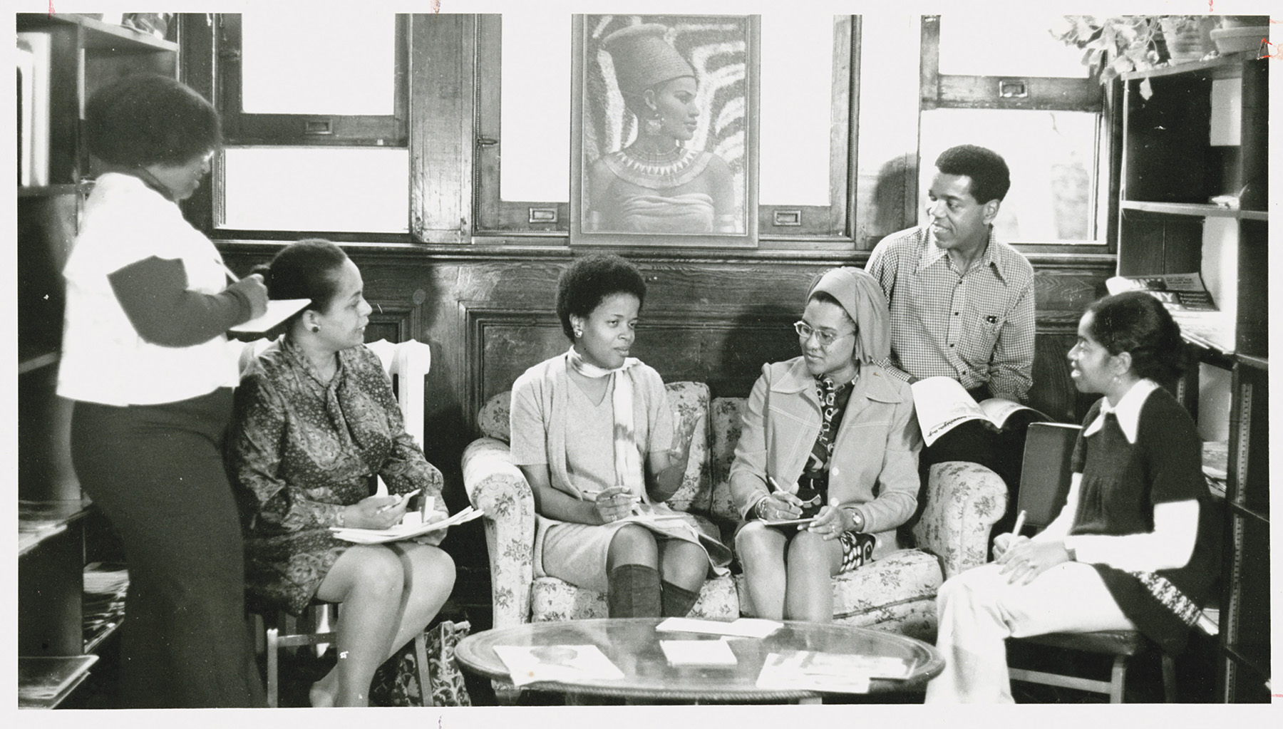 An old black-and-white photo of five people sitting on an old ornate couch in front of a coffee table while a person stands looking at them with a notepad preparing to write.