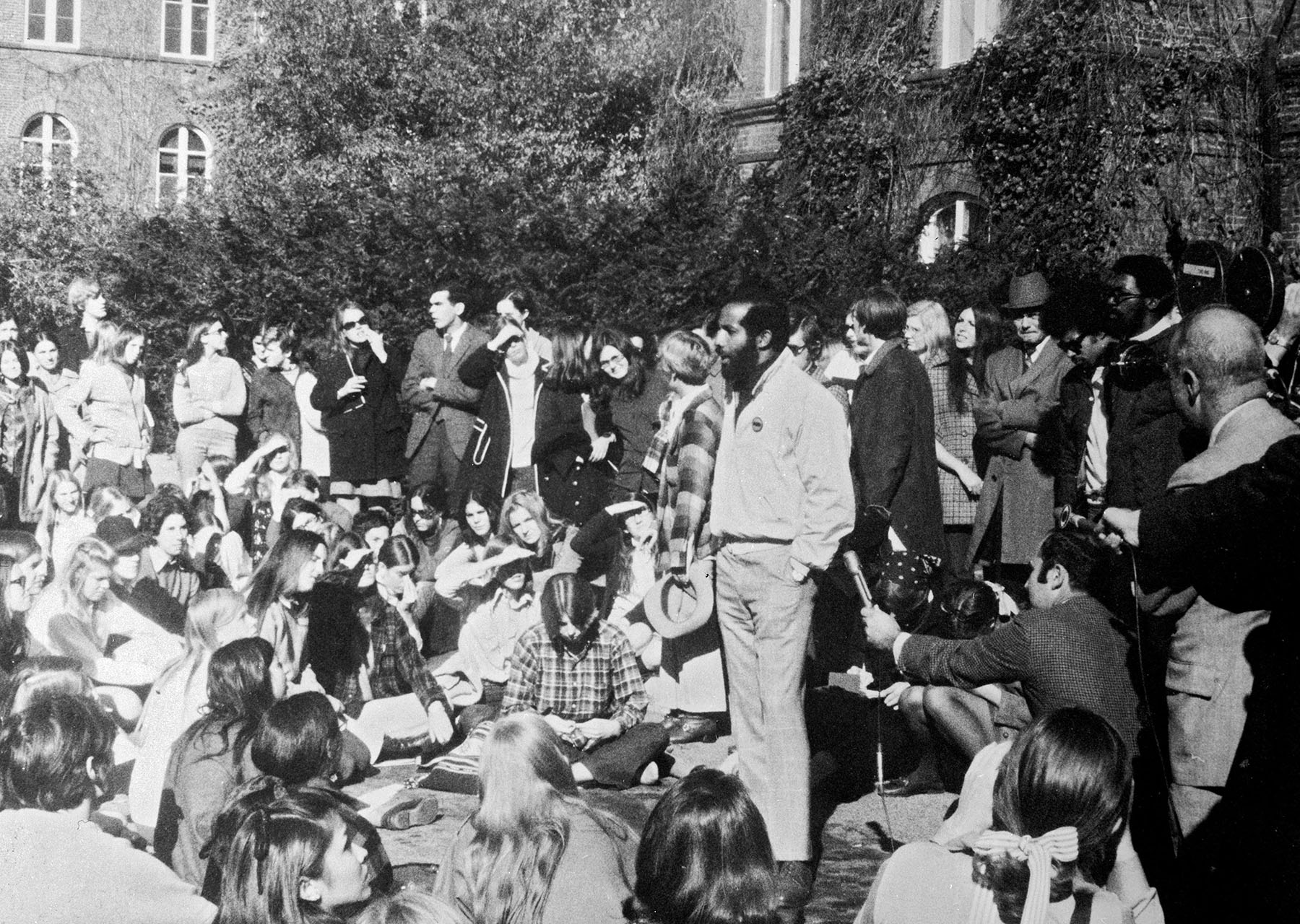 A very large group of people outside in front of a brick building (Main Building). Some people are sitting while some are standing. Prominently, there is a man in the center speaking or possibly yelling.