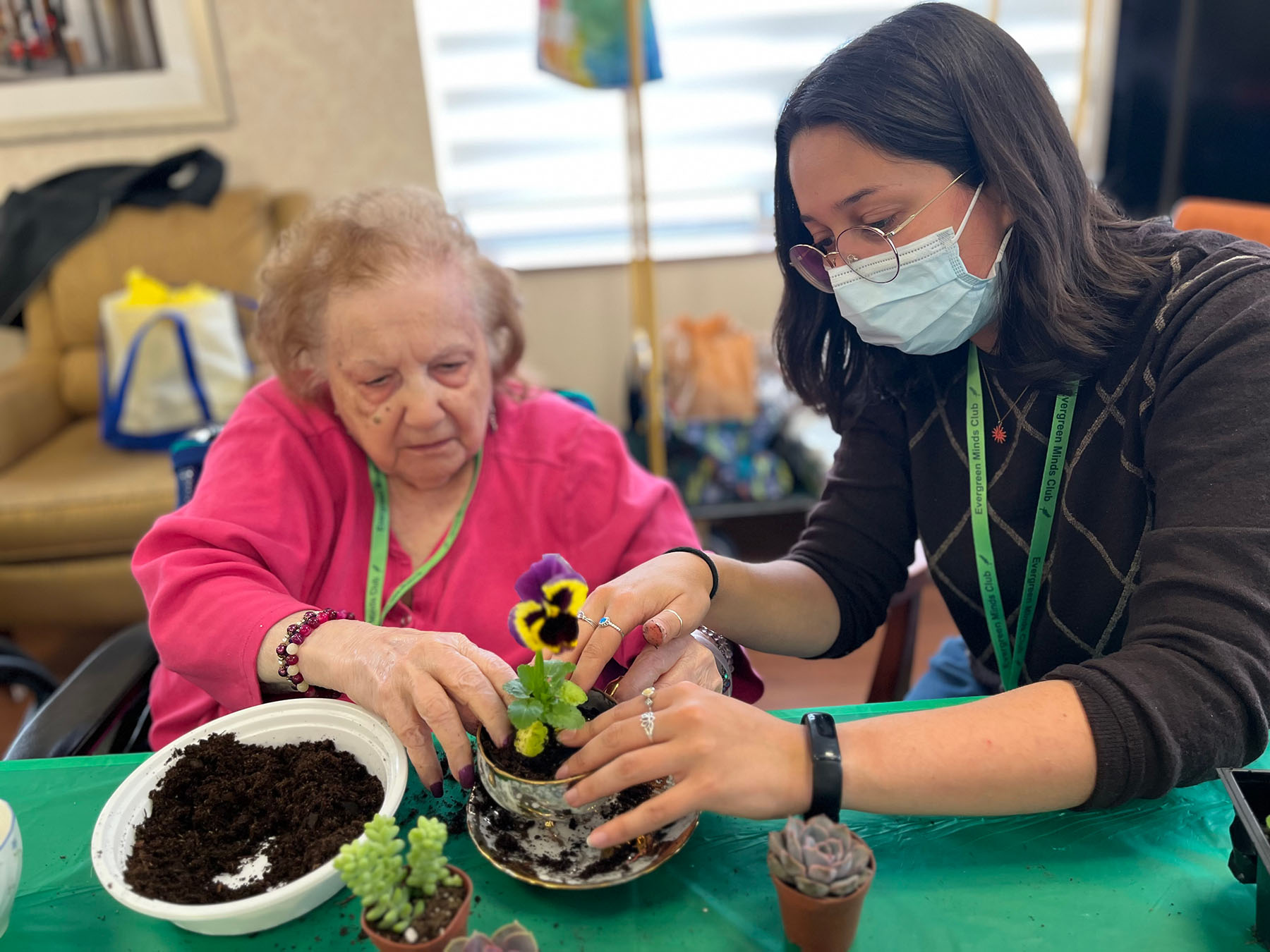 A woman in a face mask assists an elderly woman in planting a flower in a teacup.