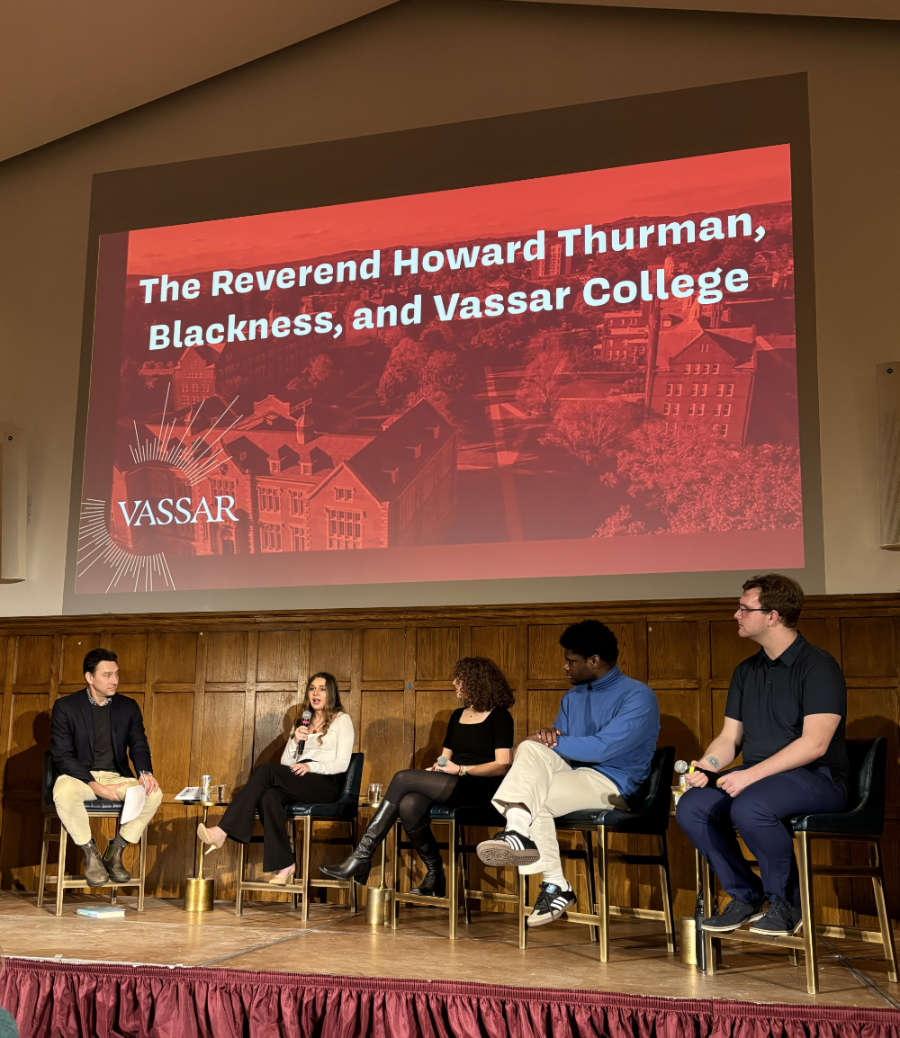 A group of students sit on a stage in front of a projected slide that reads "The Reverend Howard Thurman, Blackness, and Vassar College".