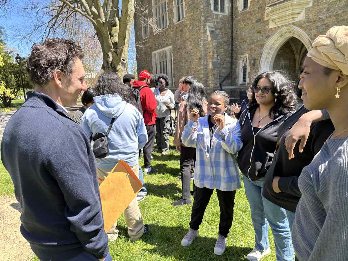 A group of people stand outside on a sunny day. Behind them is a stone building.