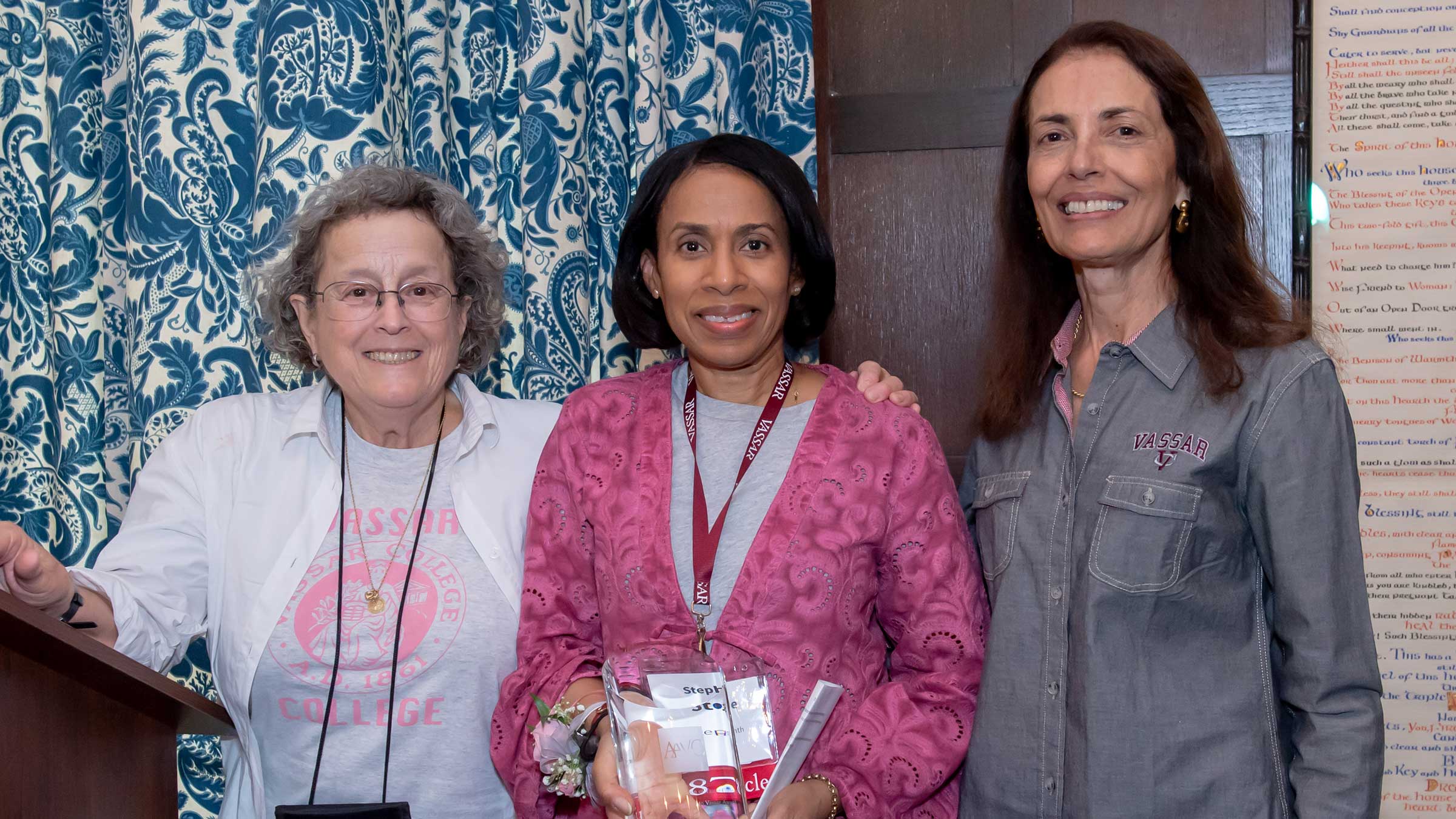 Three people smiling and standing side by side. The person in the middle is holding an award.
