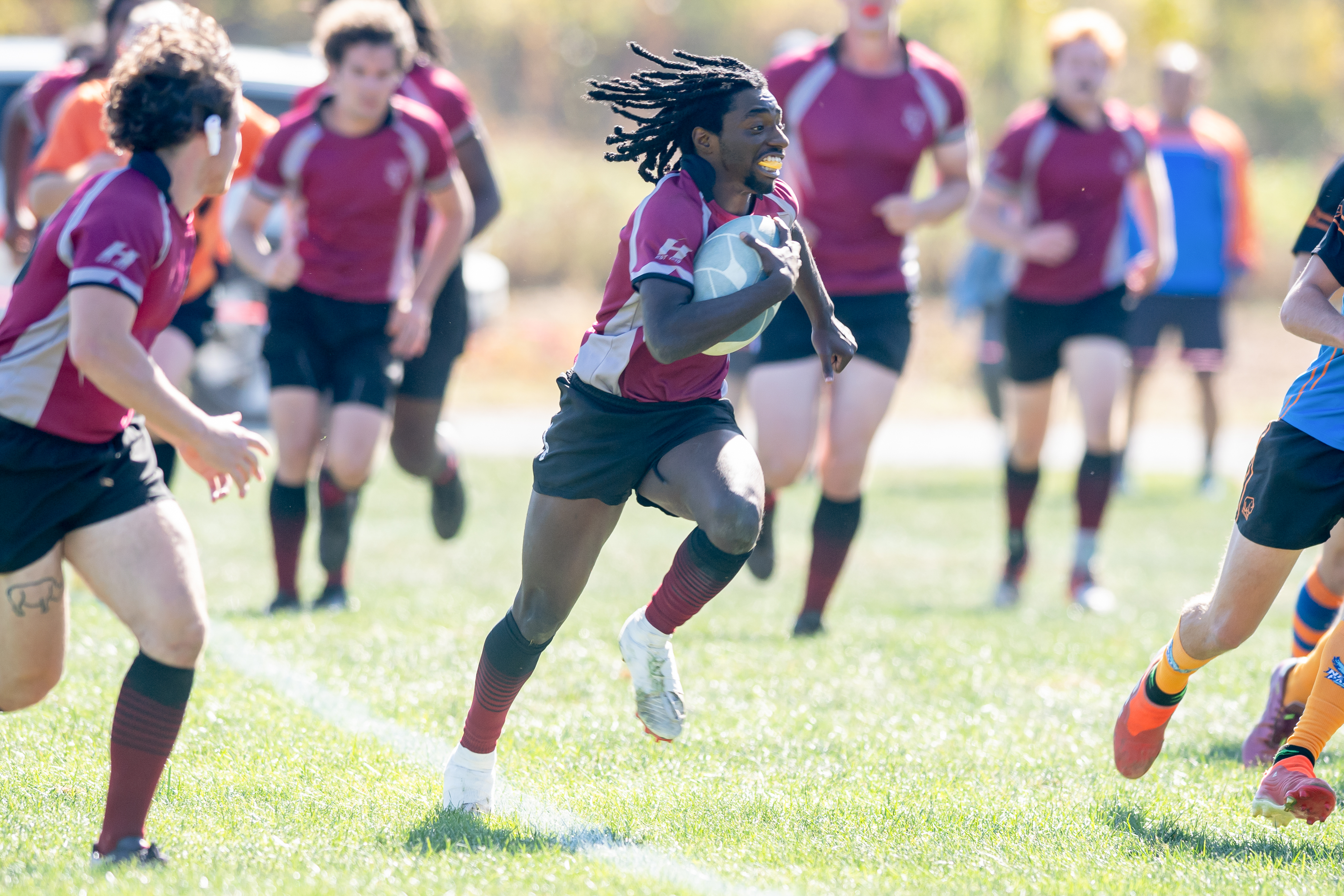 A rugby player with braids streaming behind him and a mouthguard clamped in his teeth runs with the ball during a game.