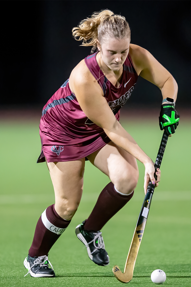 A field hockey player holding her stick with both hands bends forward to hit the ball.