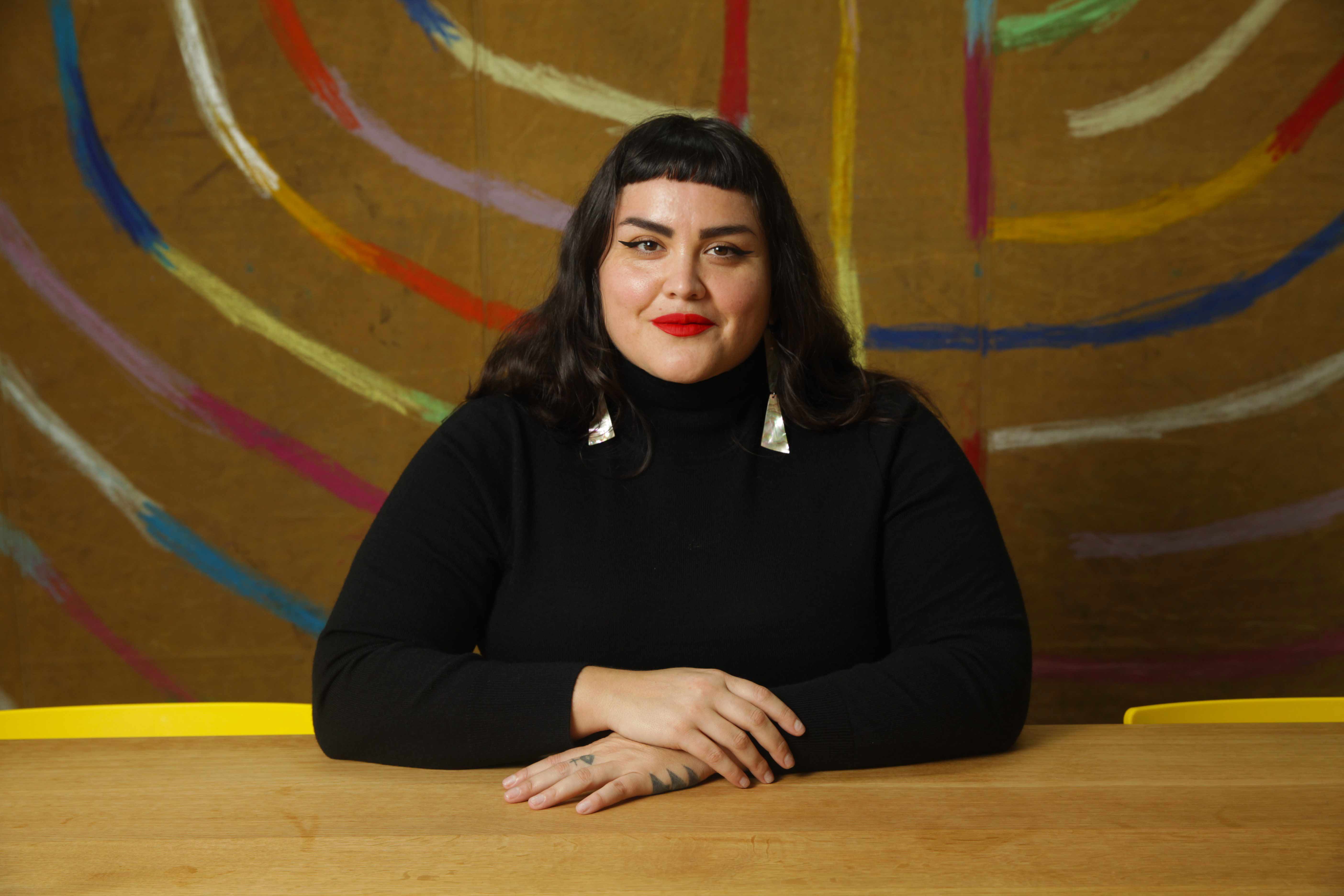 A portrait photo of Sarah Biscarra-Dilley sitting at a desk in front of a circular multi-colored painted motif.