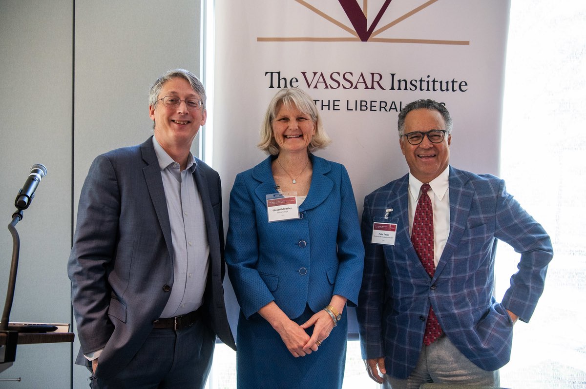 Three people stand in front of a banner that says “The Vassar Institute for the Liberal Arts”. President Elizabeth Bradley, a person with short gray hair and a blue dress, stands between a person with gray hair and a gray suit and a person with gray hair and a blue checked suit.