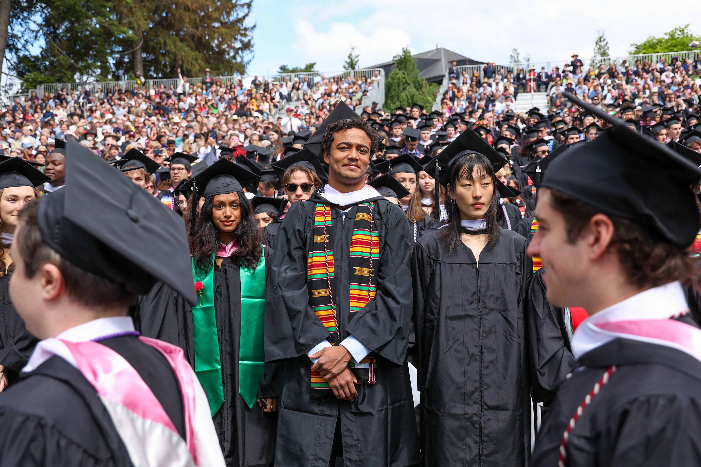 Large group of people in graduation gowns and mortar boards standing in audience.