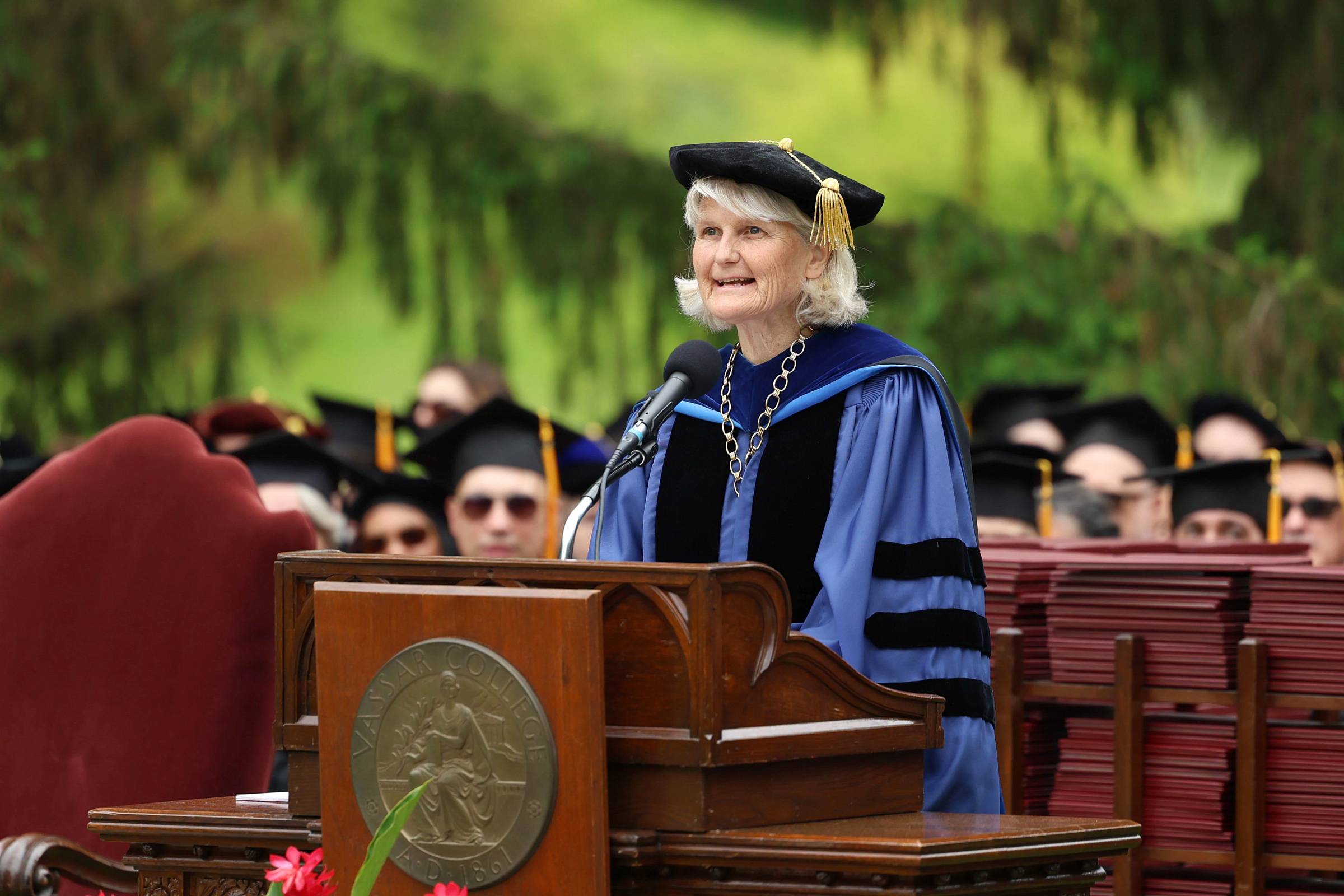Person in a graduation gown and mortar board speaking to a audience off camera.
