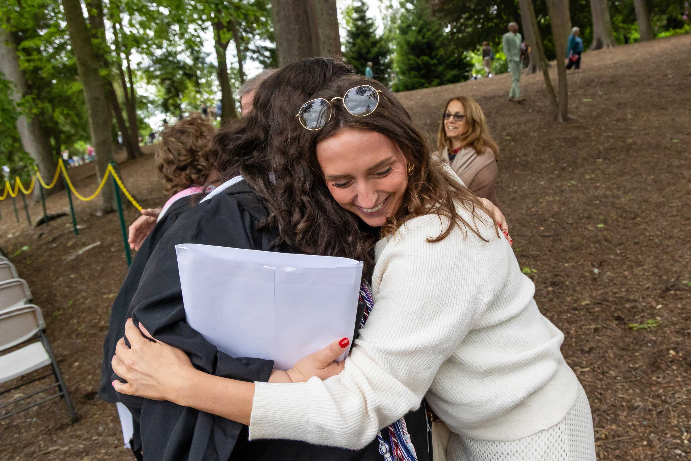 One person hugging another person wearing a graduation gown and mortarboard cap.