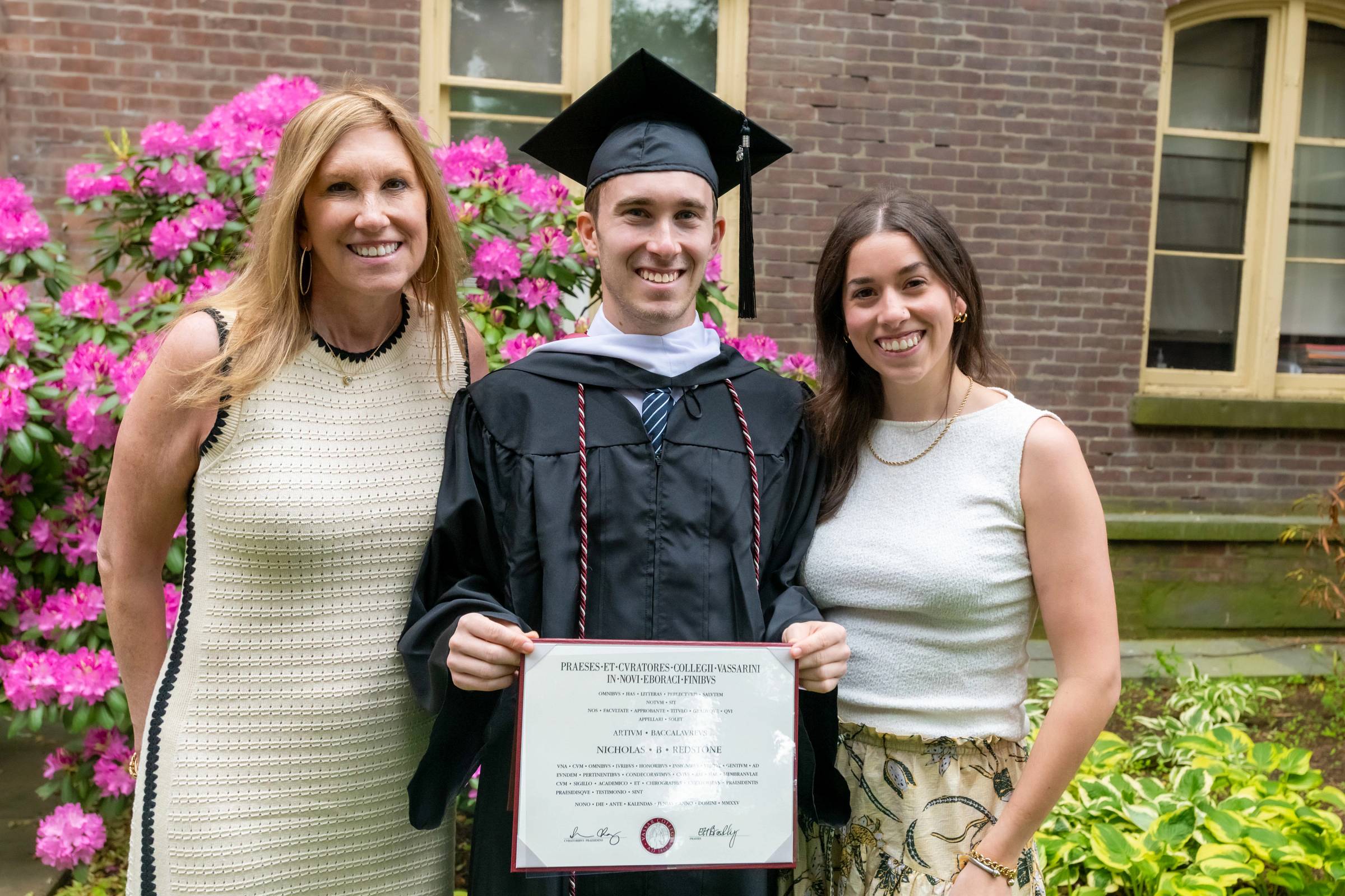 Two smiling people standing on either side of a smiling person in a graduation cap and gown holding a diploma.