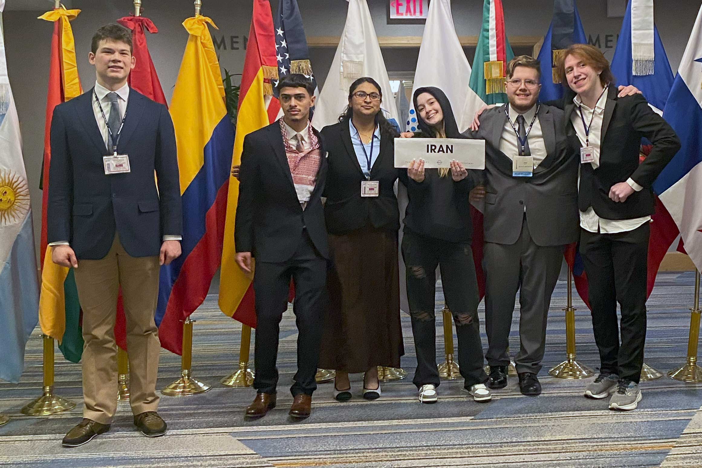 A group of smiling people standing in front flags from many different countries. A person in the middle is holding a sign that reads: Iran.