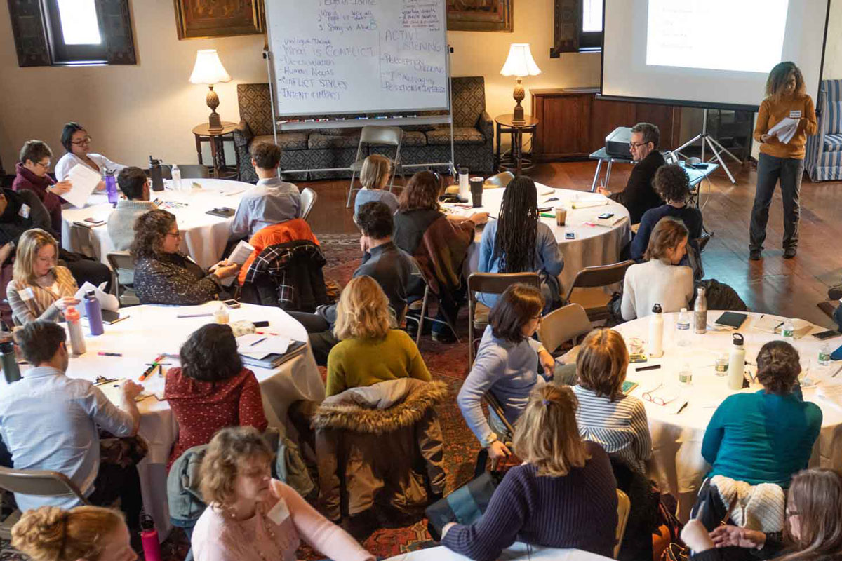 A room of people seated at round tables listening to a speaker who is standing.