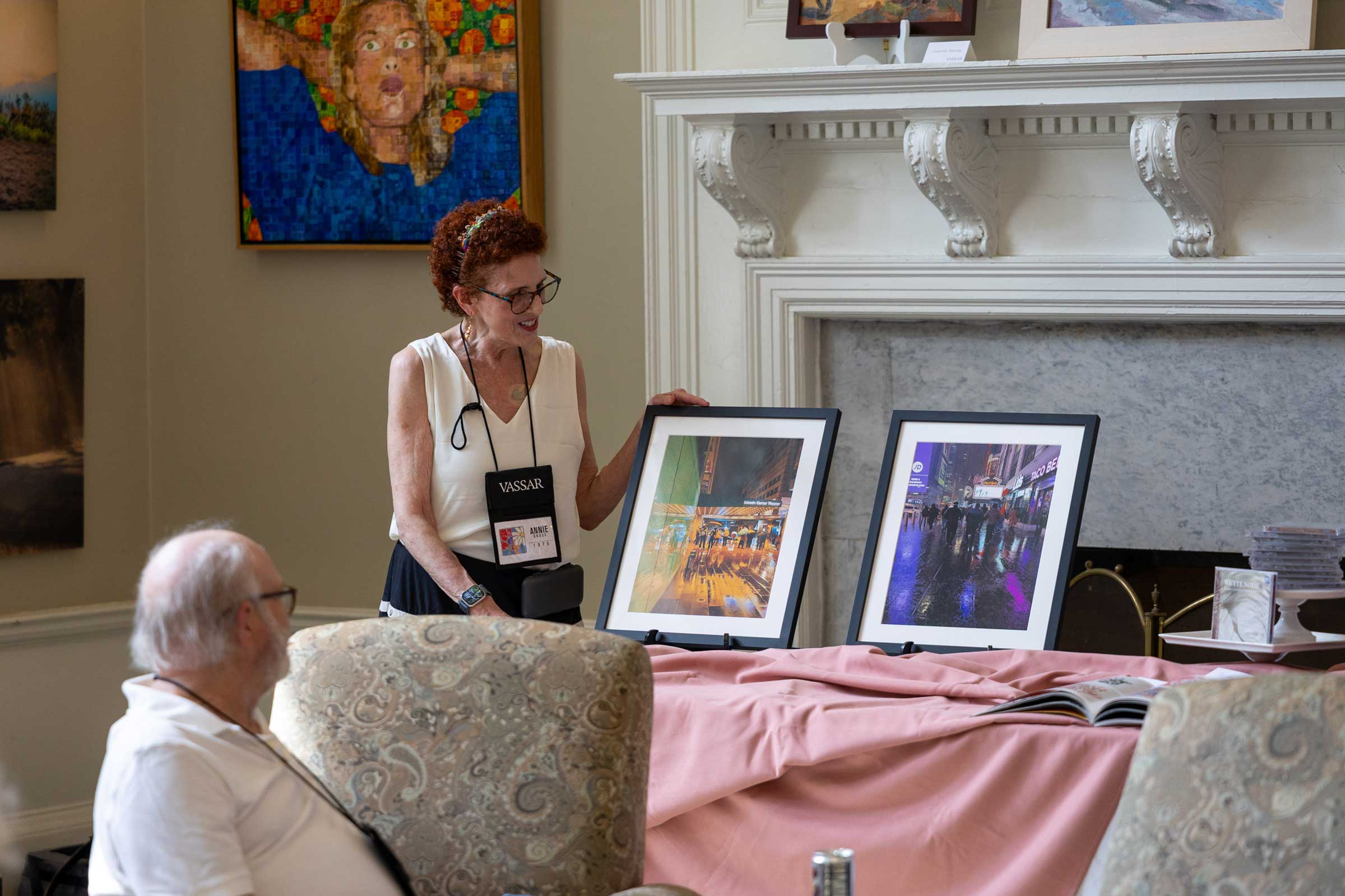 A person standing behind a table holding and artwork and talking to an audience, mostly off-screen.