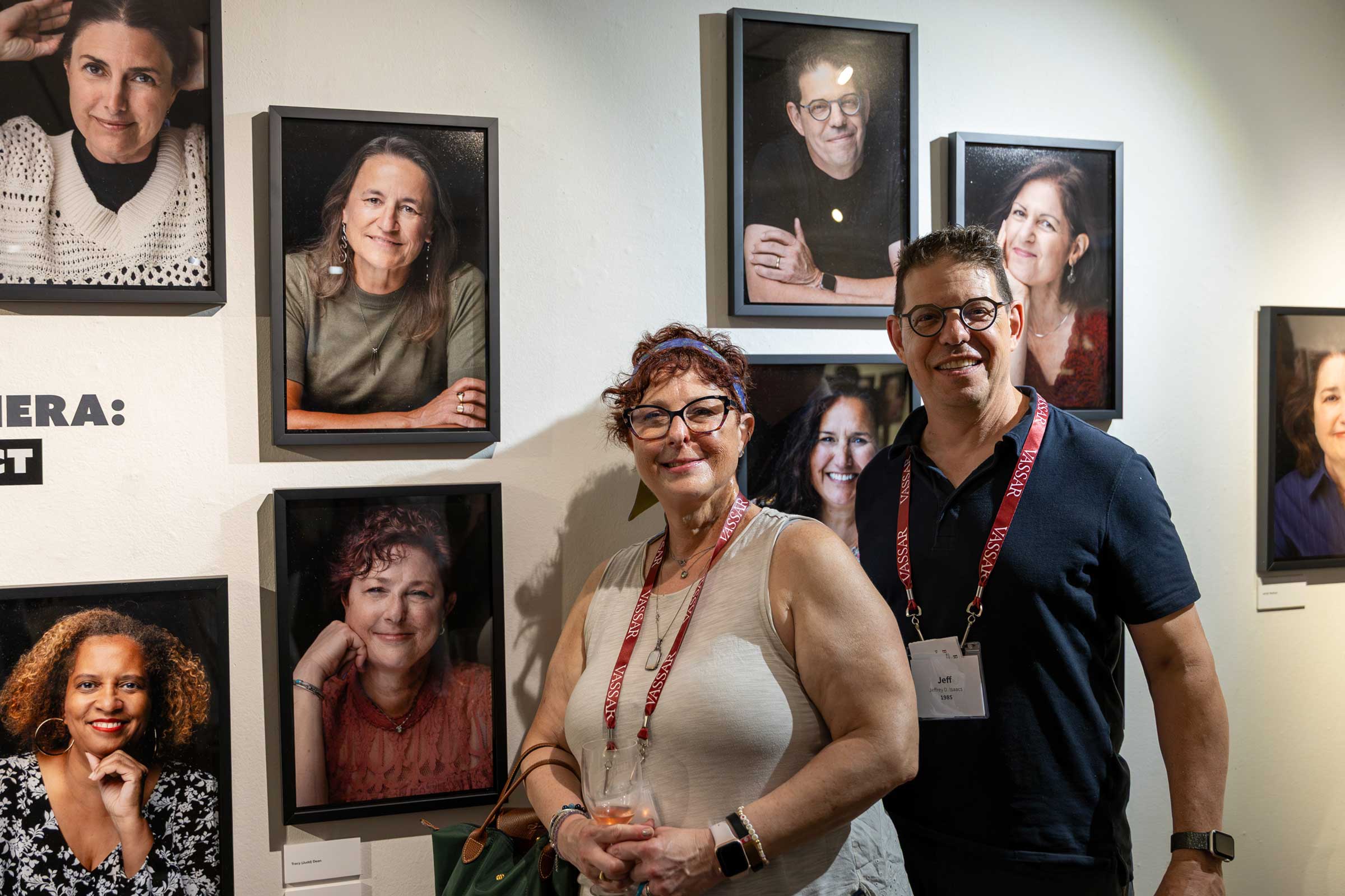 Two smiling people standing in front of a wall of photographic portraits.