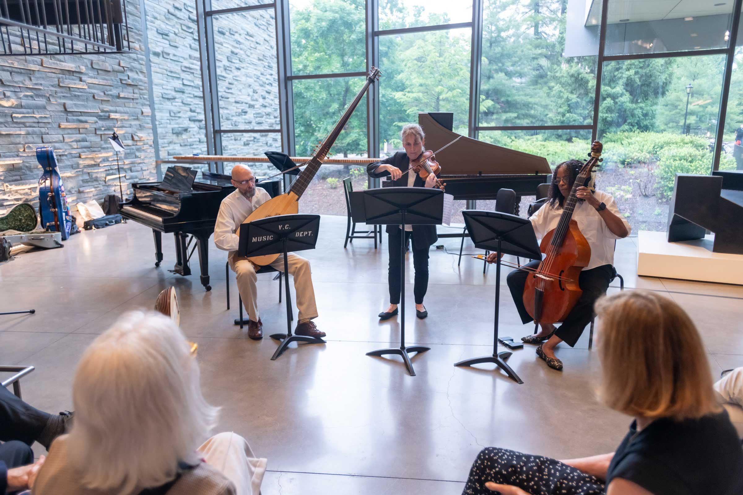 Three musicians playing string instruments standing before a seated audience.