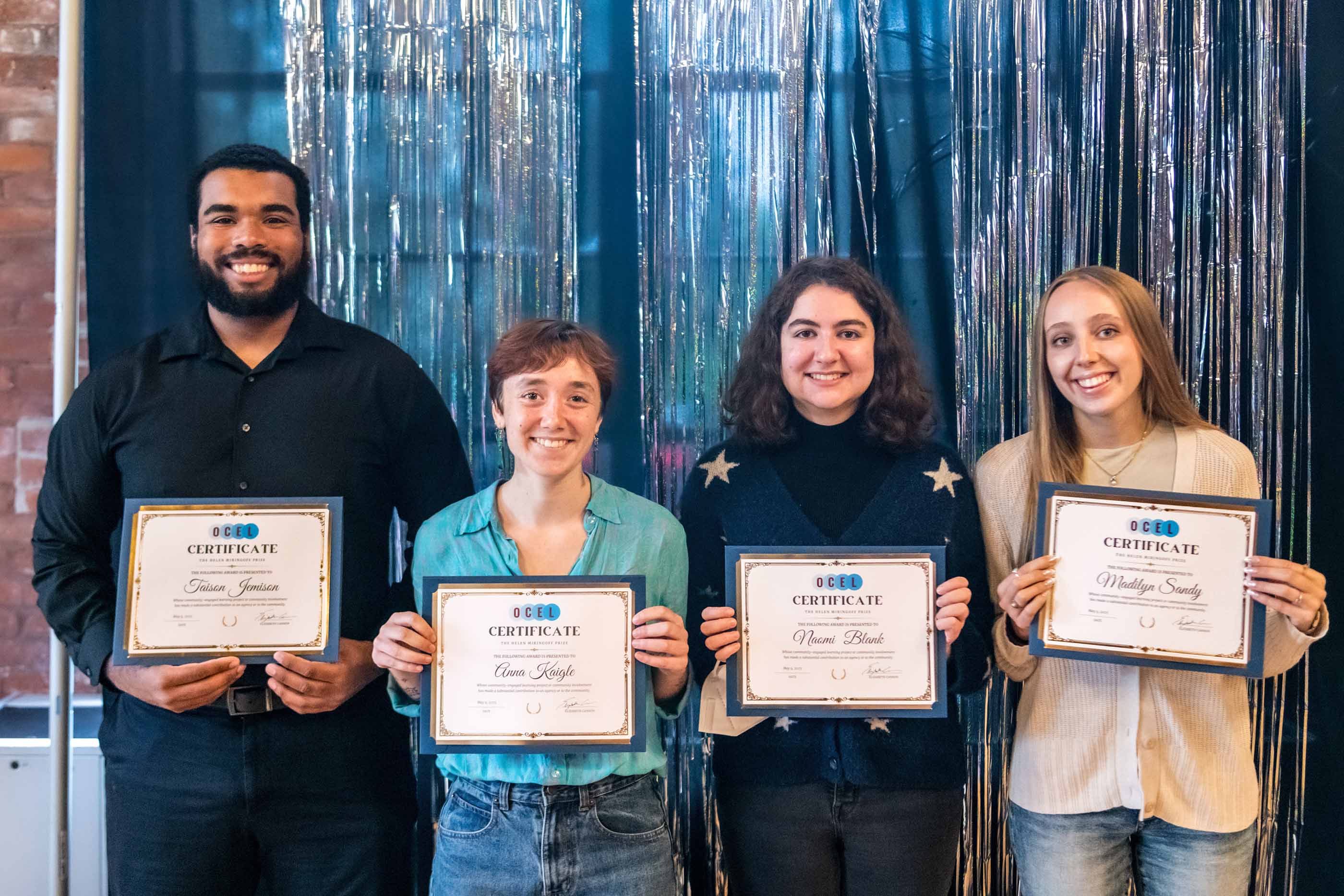 Four people stand together in front of in front of a shiny metallic streamers, smiling and holding Vassar OCEL certificates. From left to right: a person in a black shirt and pants, a person in a light blue shirt and jeans, a person in a blue shirt with stars and jeans, a person in a beige top and jeans.