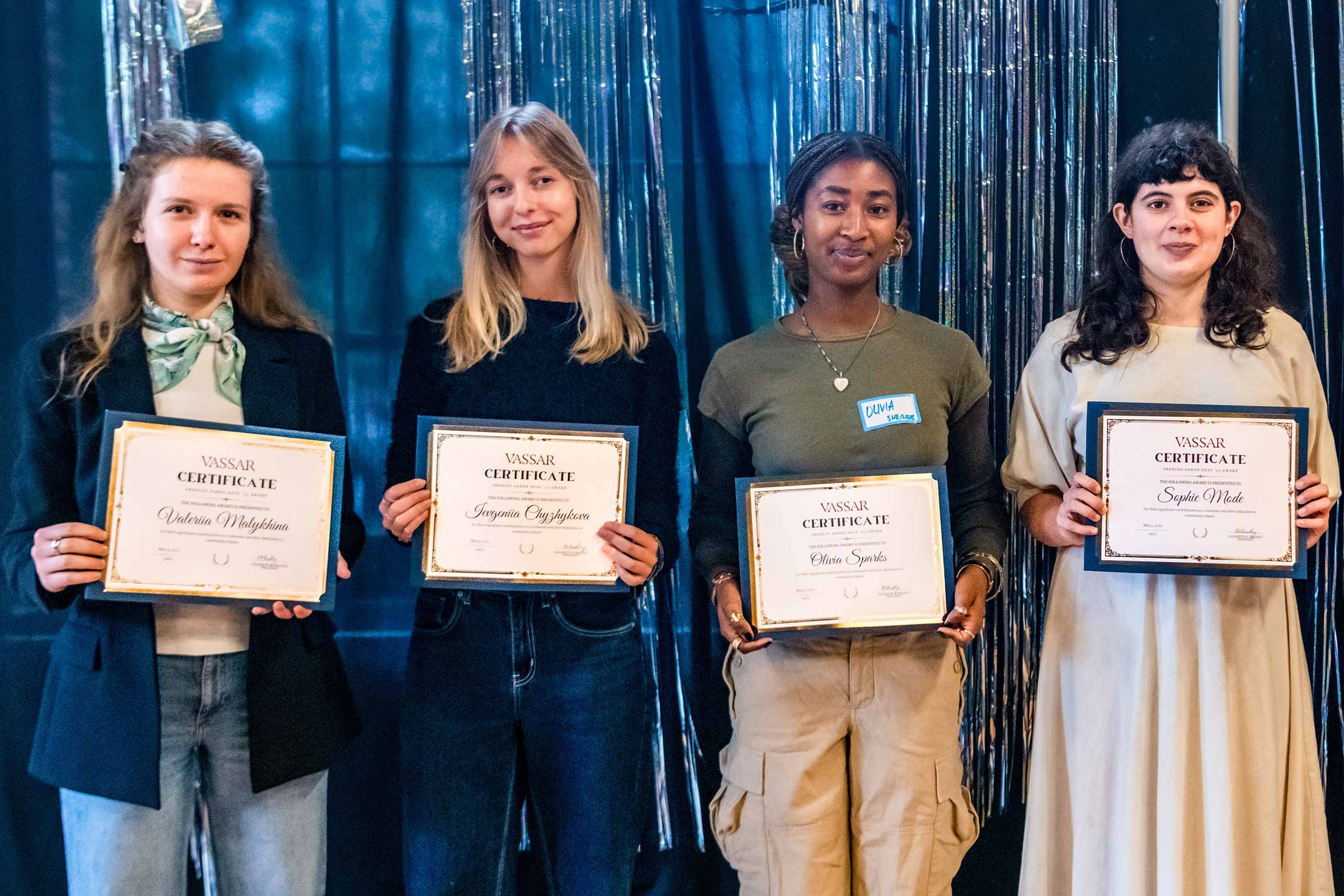Four smiling individuals stand side by side holding Vassar certificates in front of a shiny metallic streamers. From left to right: a person in a blue jacket, white shirt and jeans, a person in a blue shirt and jeans, a person in a olive shirt and tan pants, and a person in a beige dress.
