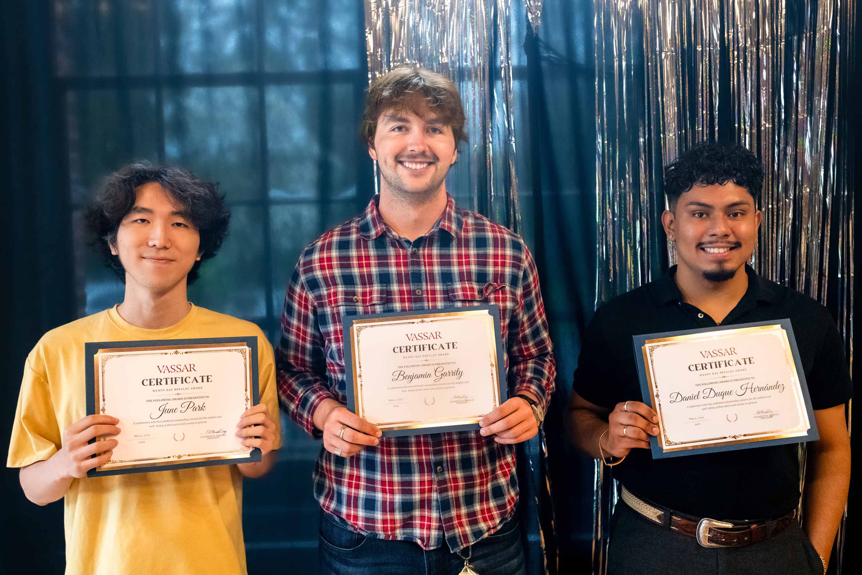Three smiling individuals stand side by side holding Vassar certificates in front of a shiny metallic streamers. The person on the left wears a yellow shirt, the middle person wears a red and blue plaid shirt, and the person on the right wears a black shirt.