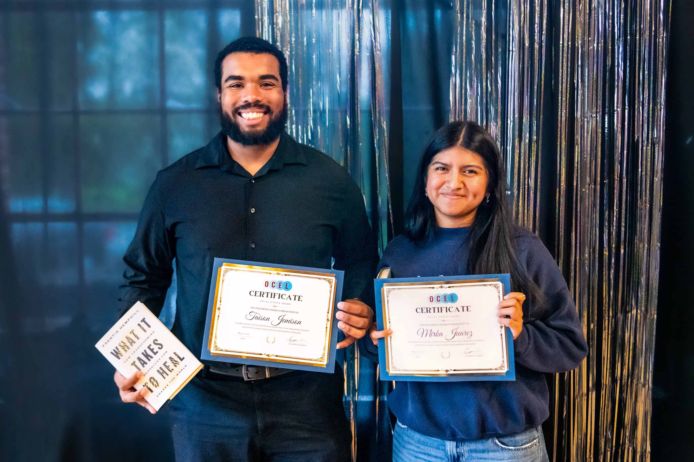 Two smiling individuals stand side by side holding certificates from OCEL. The person on the left also holds a book titled ‘What It Takes to Heal.’ They are standing in front of a dark, decorated backdrop with shiny metallic streamers.