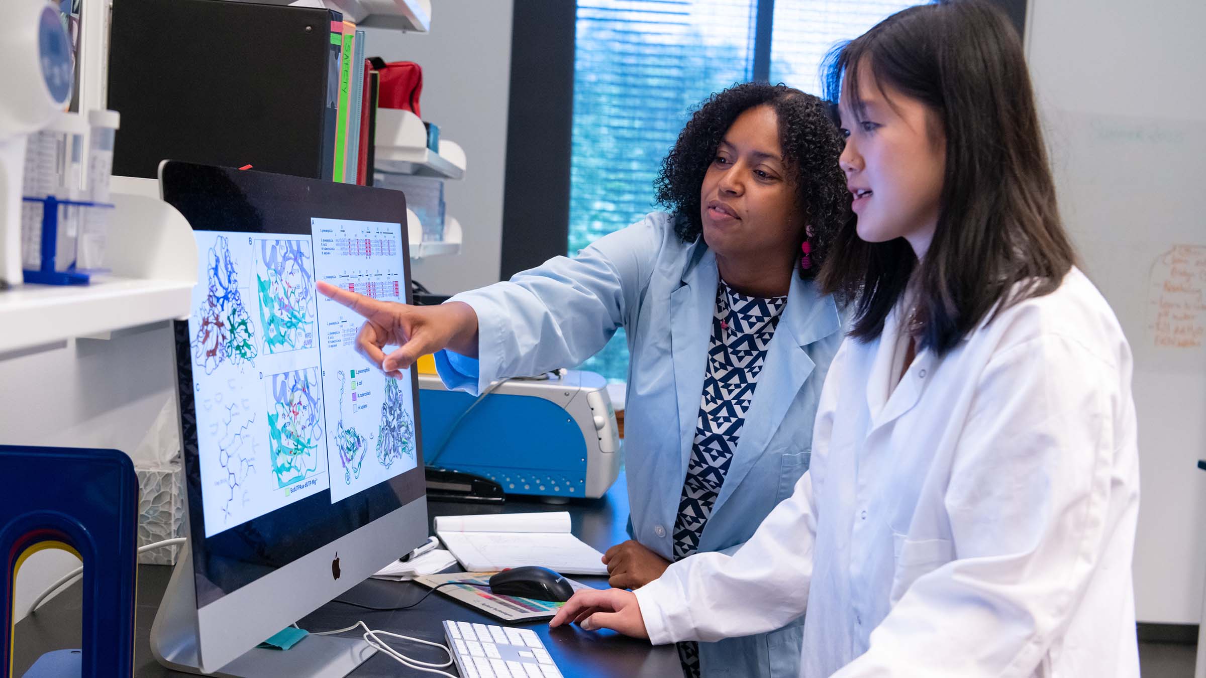 Two people wearing lab coats examine 3D protein structure models on a large computer monitor in a laboratory. One person points at the screen while the other operates the computer.