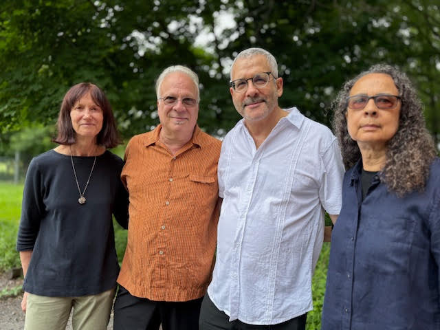 Group portrait of four people standing side by side on a green lawn, casually dressed, with trees in the background.