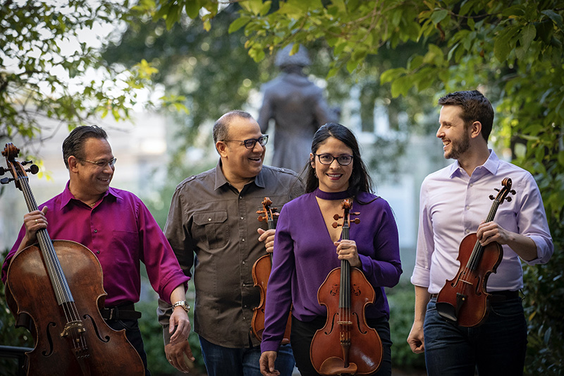 Four adults standing outdoors, posing together in front of leafy trees, smiling or looking at the camera each holding their instrument.