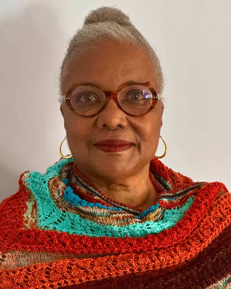 A person with short gray hair, glasses, and an orange shawl looks at the camera.