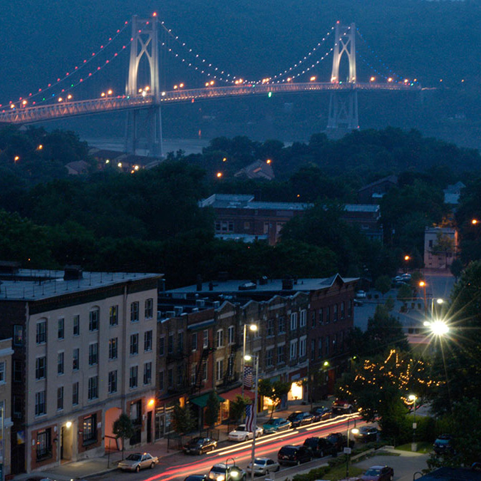 Night time view of a city block with a lighted bridge in the background.