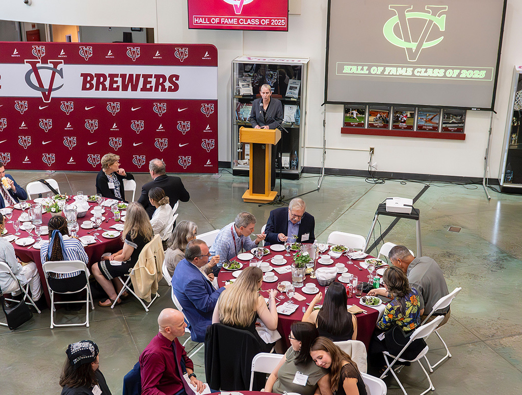A speaker at the podium welcomes an audience at round tables during a banquet.