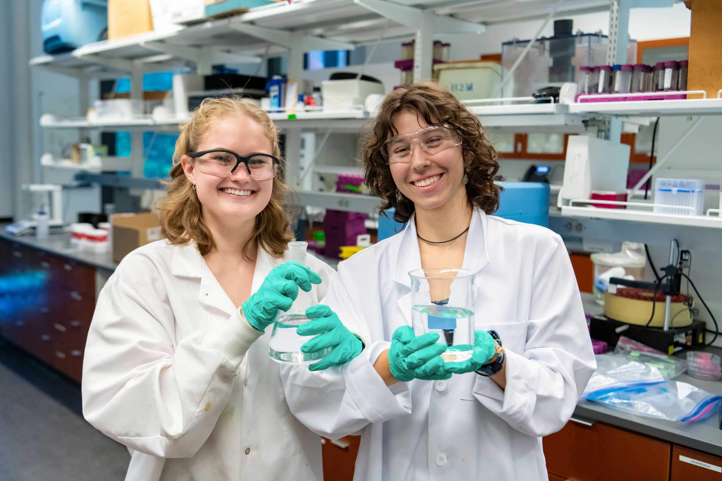 Two smiling people in a lab wearing gloves and holding lab containers.