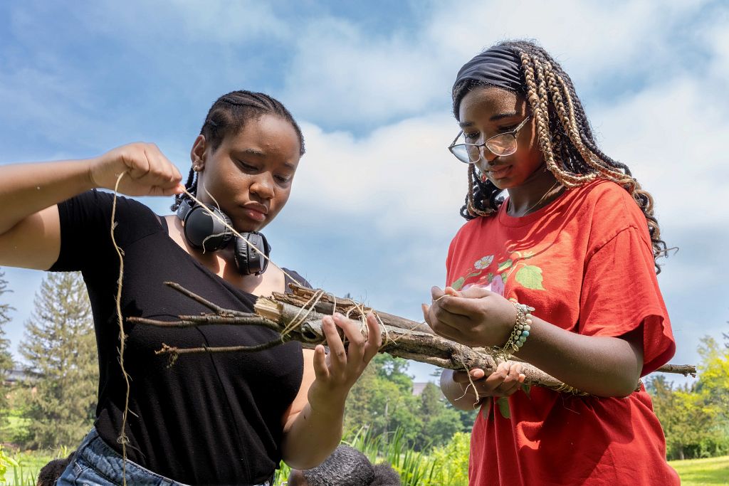A close-up, low-angle shot of two students collaborating on a project outdoors. The student on the left holds a bundle of twigs. The student on the right wraps a piece of twine around the bundle. 