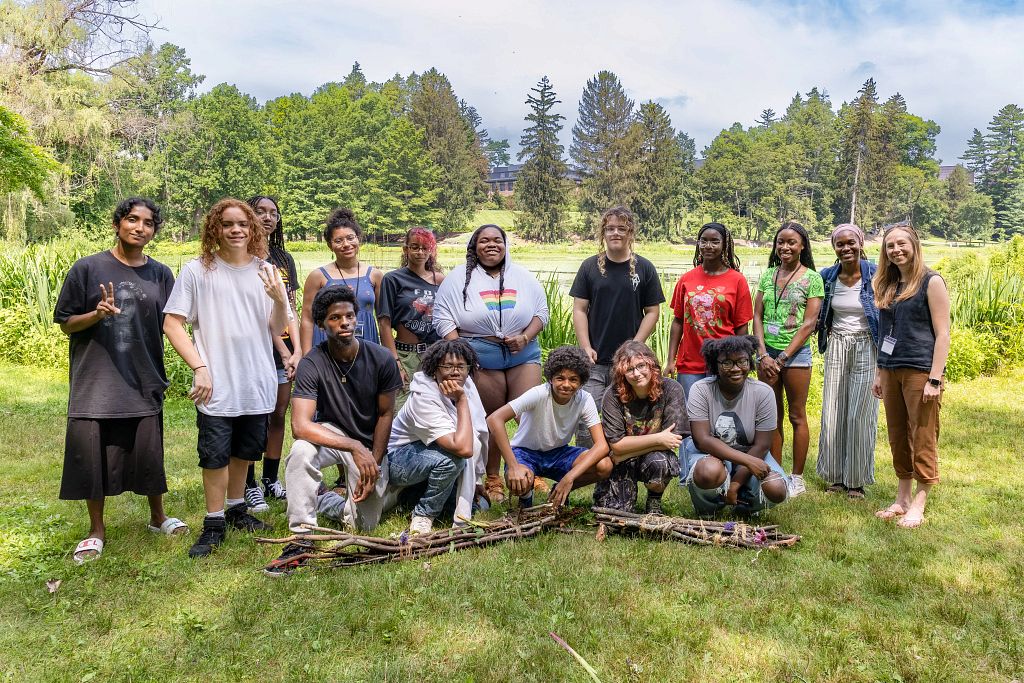 A diverse group of fifteen people is posing for a photo on a grassy lawn with a pond and a college building visible in the background.