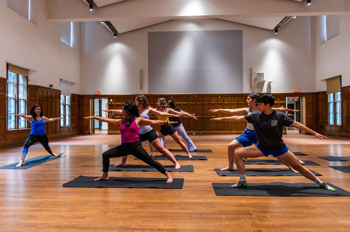 A group of people practice yoga in a spacious room with wood-paneled walls and high windows. They are arranged in rows on black mats, holding a warrior pose with arms extended and knees bent. The room has warm overhead lighting and a polished wooden floor. An instructor stands at the front, demonstrating the posture.