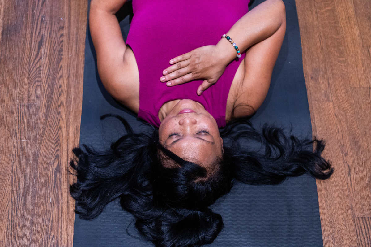 A person lies on a yoga mat with eyes closed, wearing a magenta sleeveless top. Long black hair is spread across the mat, and one hand rests gently on the chest. The black mat contrasts with the wooden floor beneath. The image shows a calm resting posture at the end of practice.