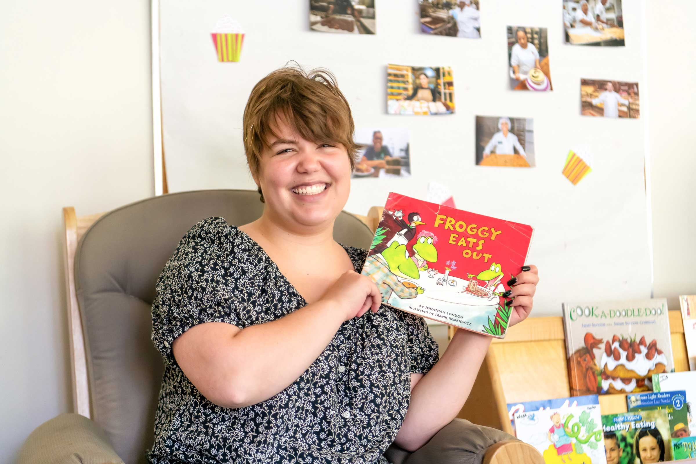 A seated person smiling and holding up a book with a title that reads, "Froggy Eats Out."