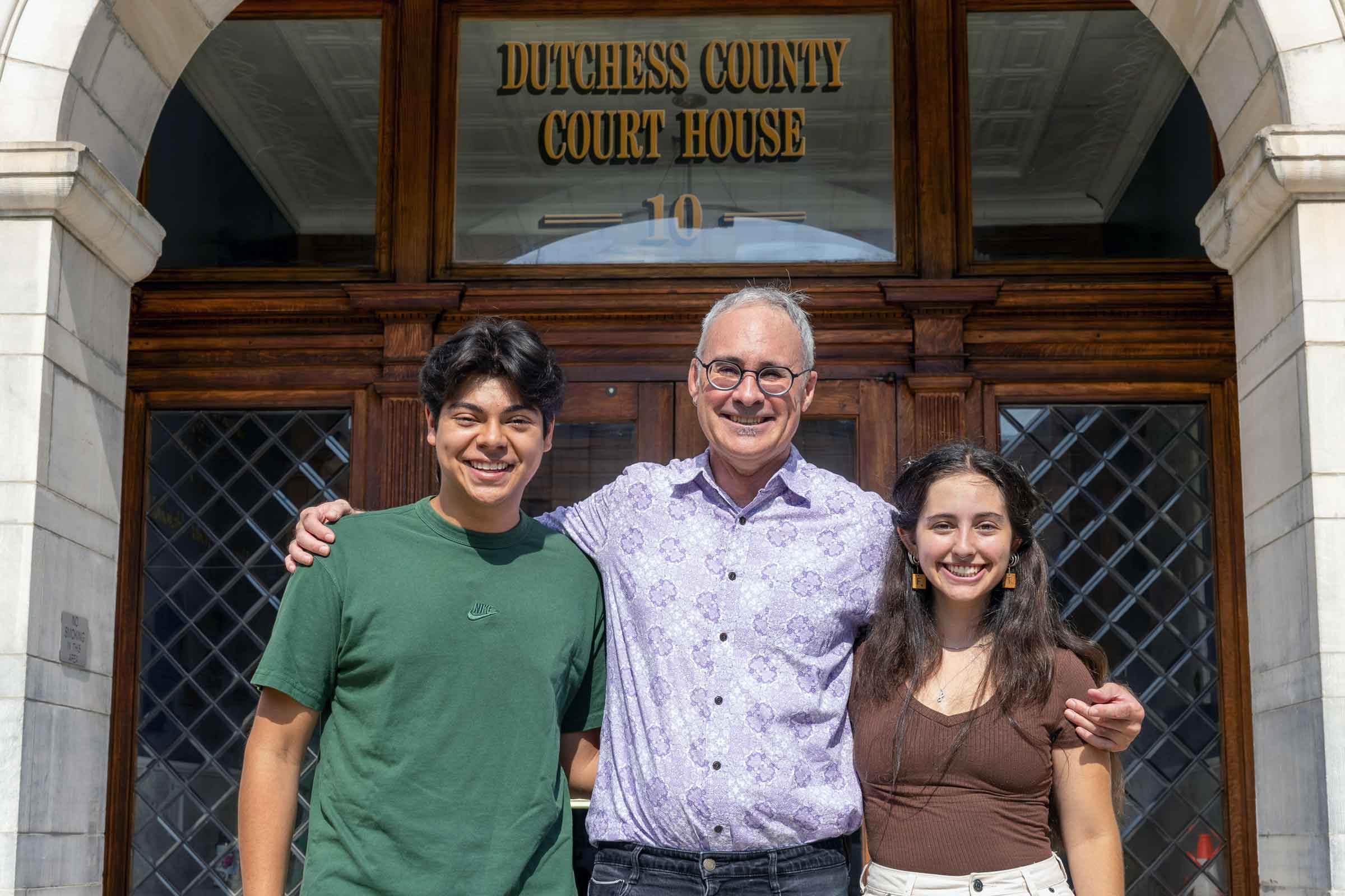 Three people standing side by side and arm in arm in front of the Dutchess County, NY courthouse.