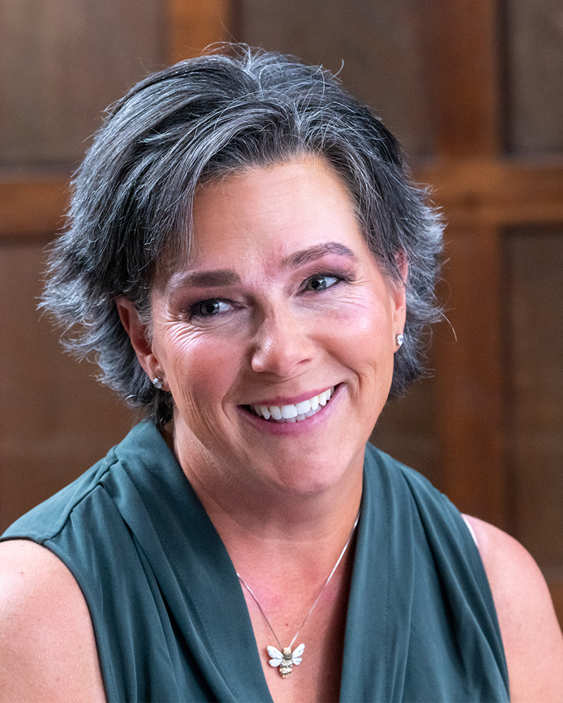 Closeup of a smiling person who has short hair and is wearing a sleeveless top and bumblebee necklace.