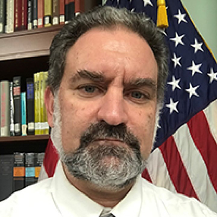 A photo of Clifford Rogers, a person with dark hair and beard and a white shirt. They are sitting in front of a bookshelf, next to an American flag.