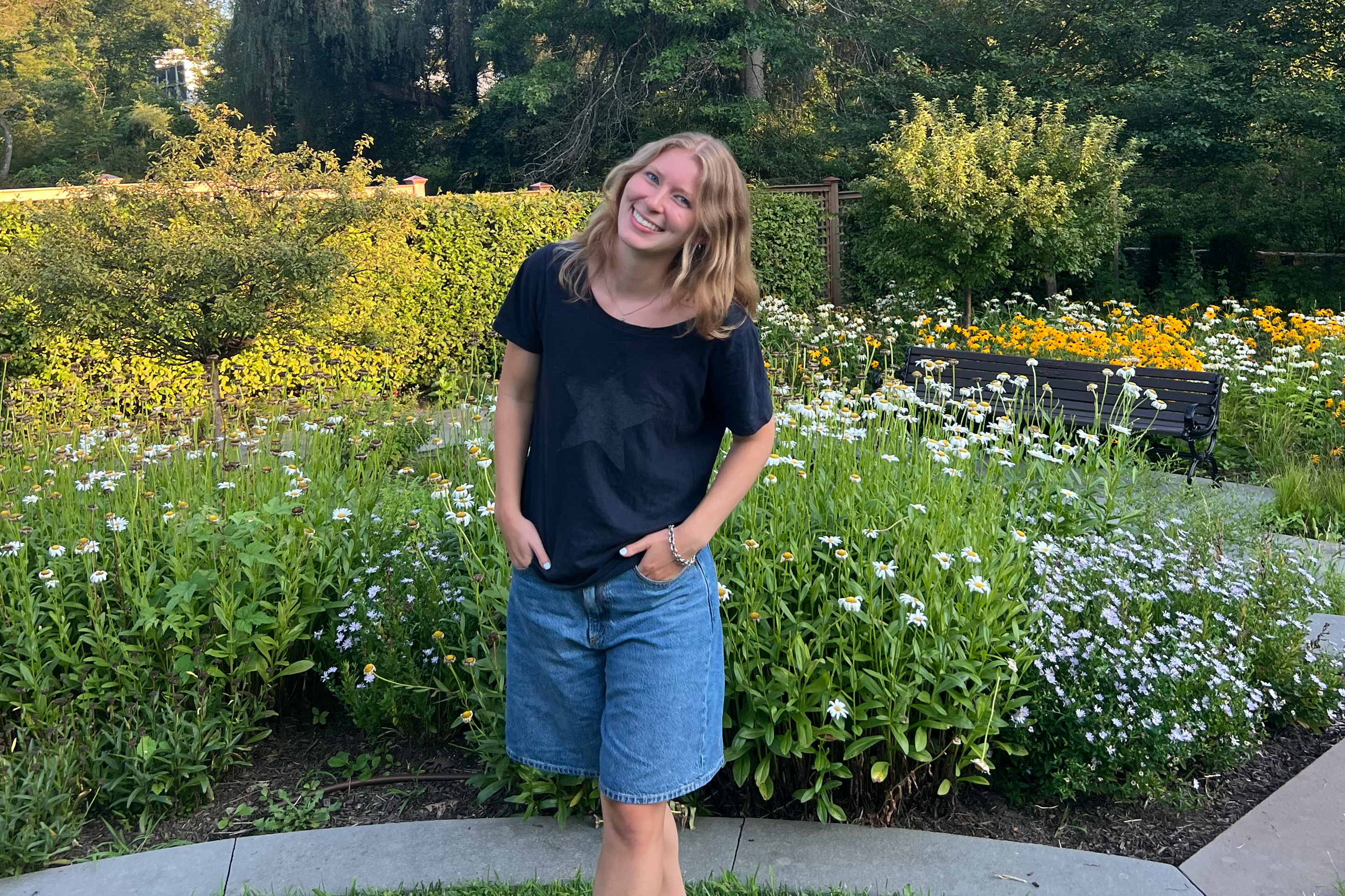 A smiling person with long hair wearing shorts and a T-shirt stands in front of a garden full of daisies.