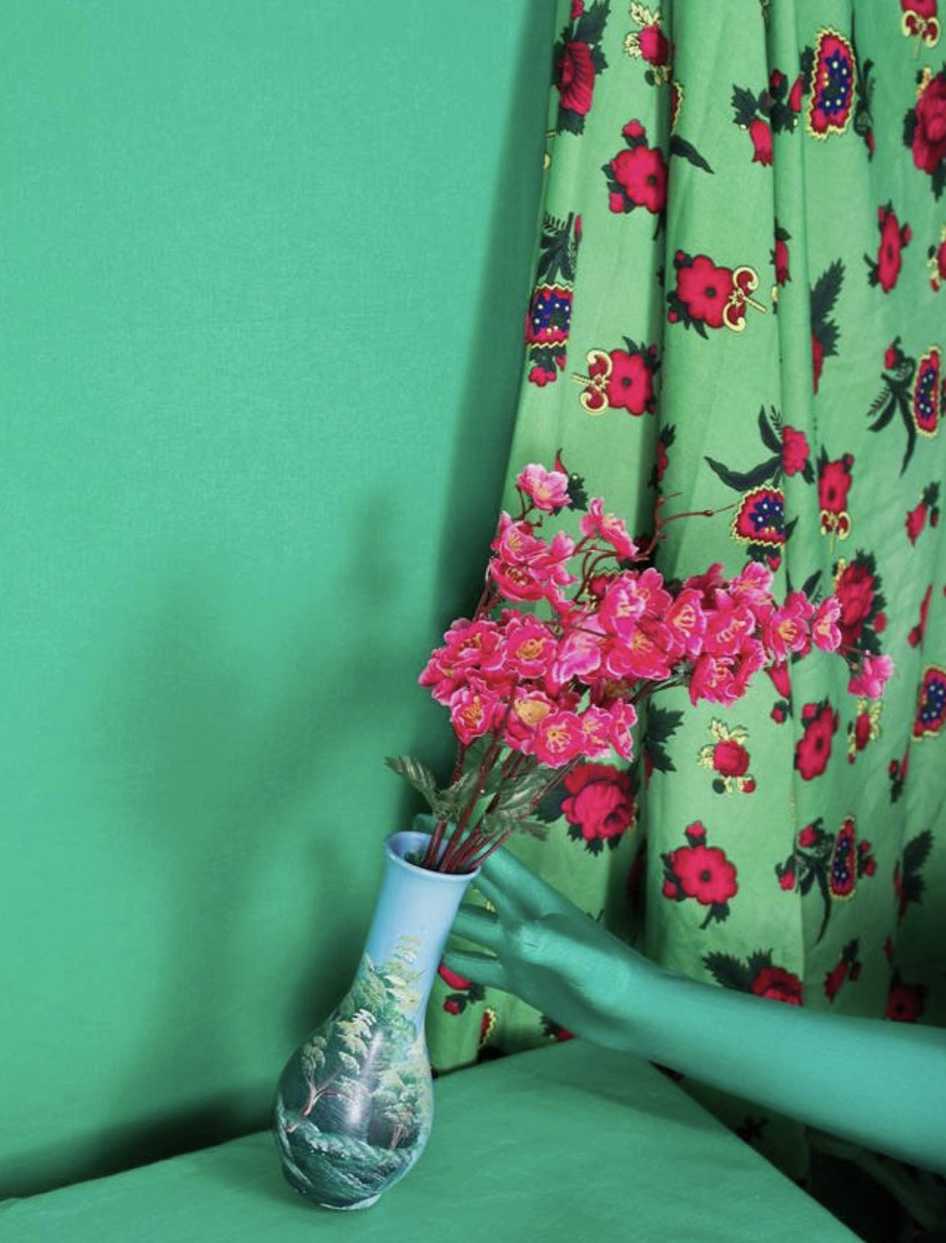 A photo of a green room, with the plastic hand of a dummy reaching for a vase with pink flowers in it.