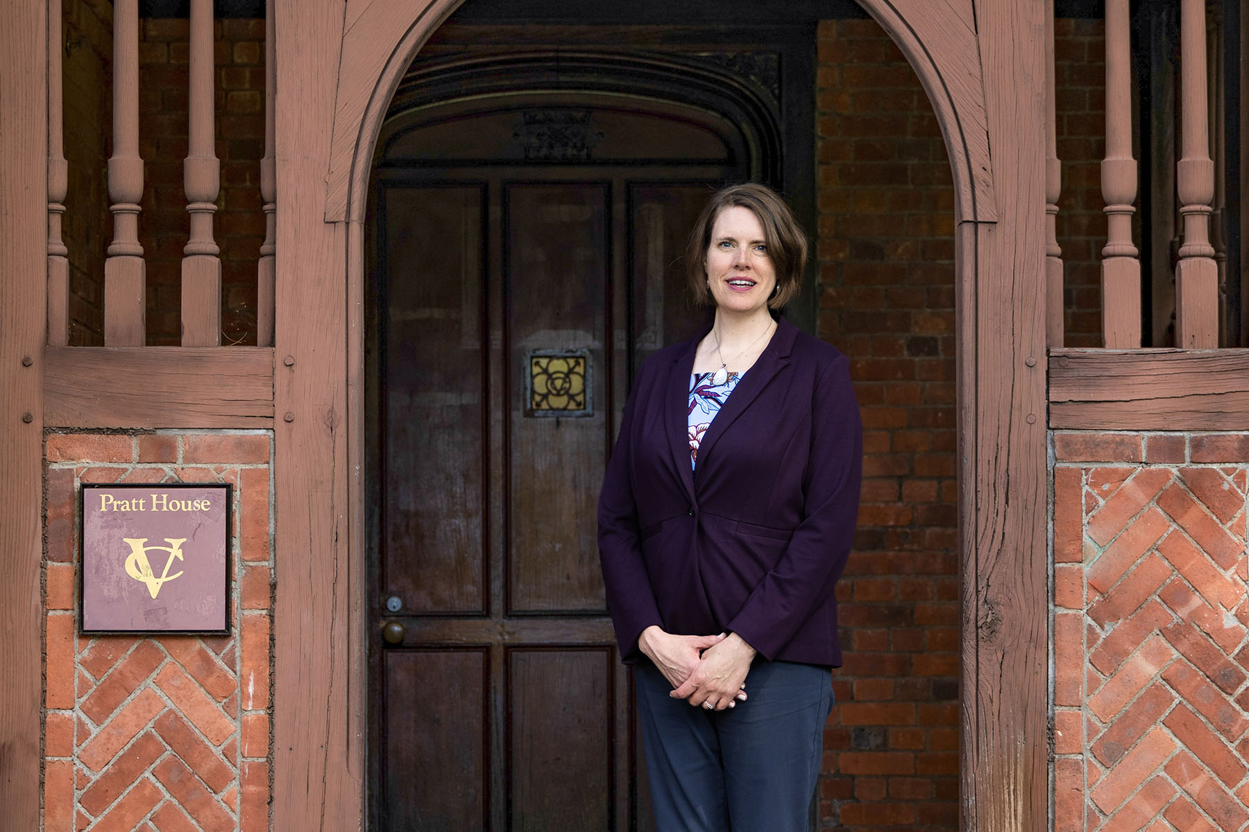Callista Isabelle in a purple blazer stands in front of a wooden doorway at Pratt House, with a sign displaying the Vassar College logo on the brick wall beside her.
