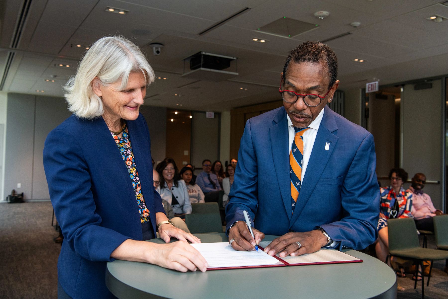 Two people signing a document at a table, with others seated in the background.
