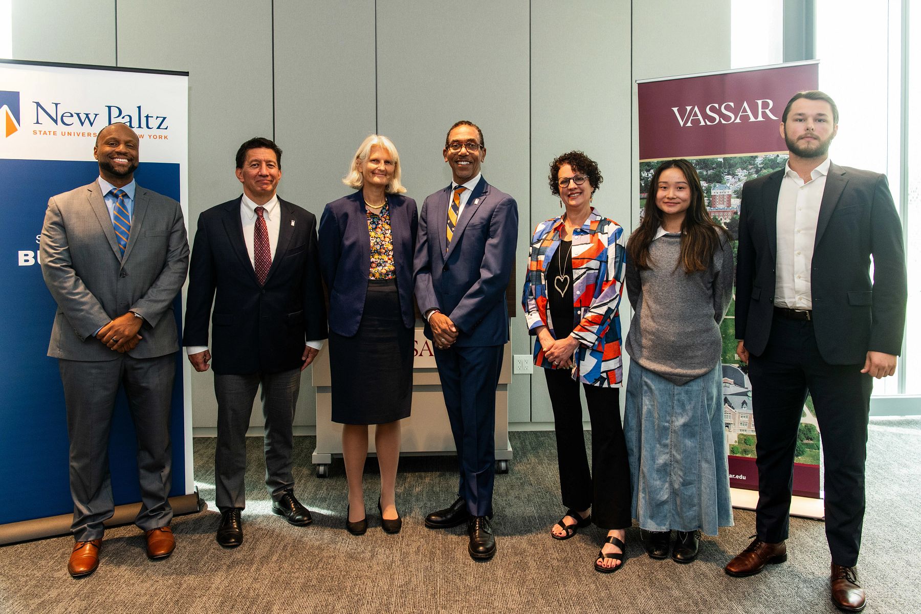 Seven people standing side by side in front of Vassar and SUNY New Paltz banners.